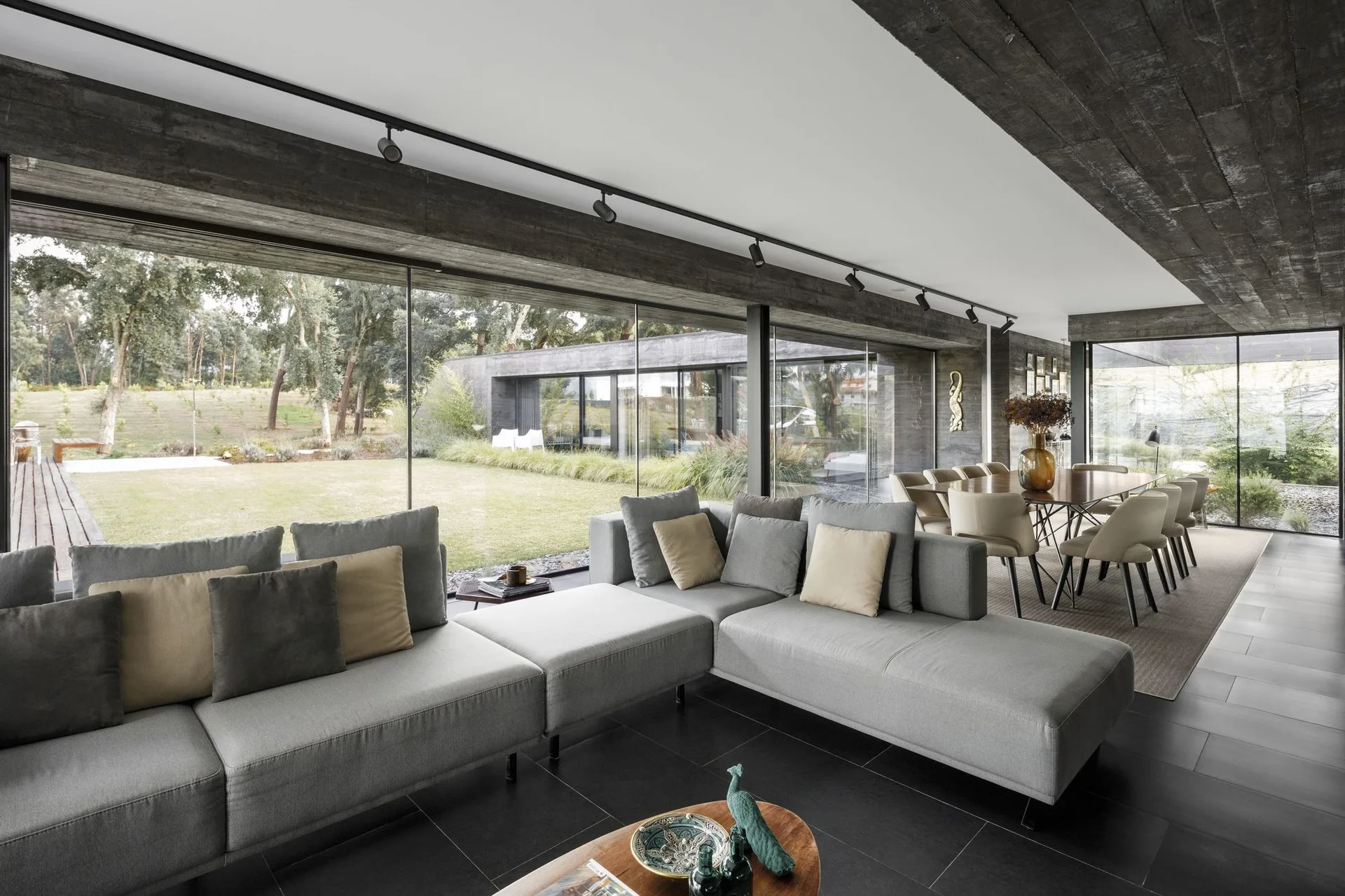 A modern open-plan living room with a grey modular sectional sofa, concrete ceilings, and floor-to-ceiling glass doors opening to a grassy lawn.
