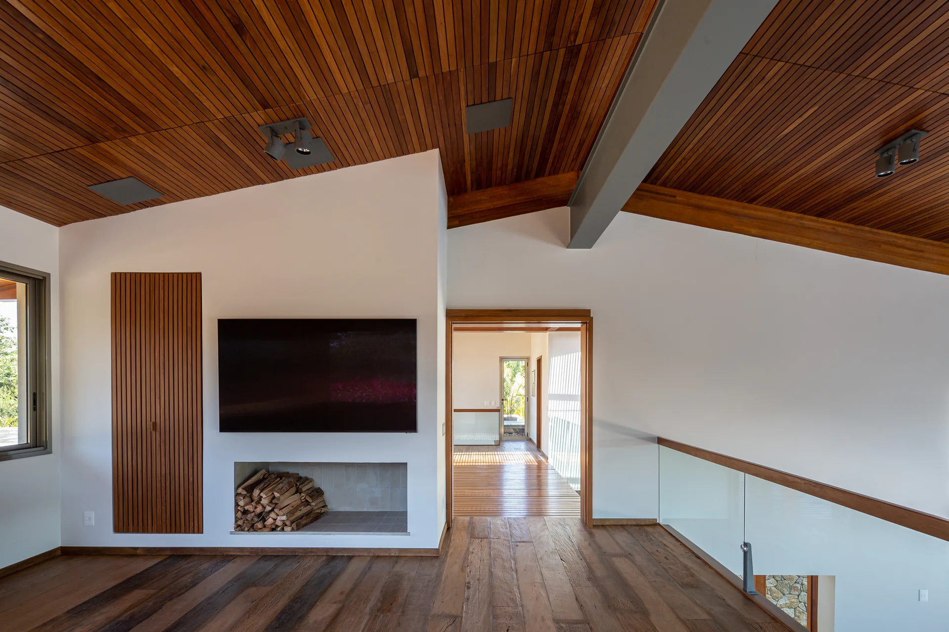 A contemporary hallway landing features a slatted wood ceiling, dark wood floors, a wall-mounted TV over a firewood niche, and a glass railing.