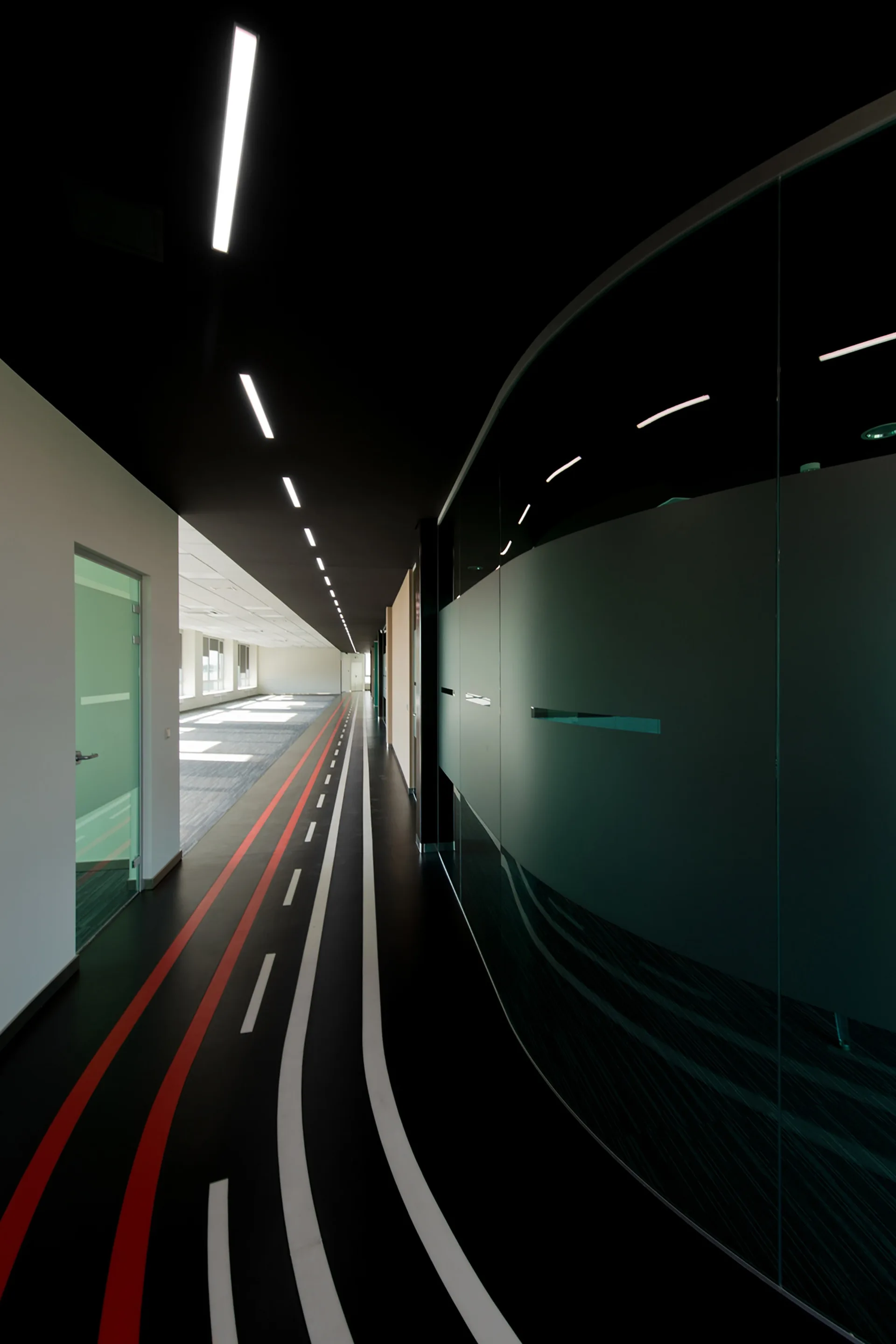 A long black office hallway features a curved glass wall partition, linear ceiling lights, and red and white racing stripes on the floor.