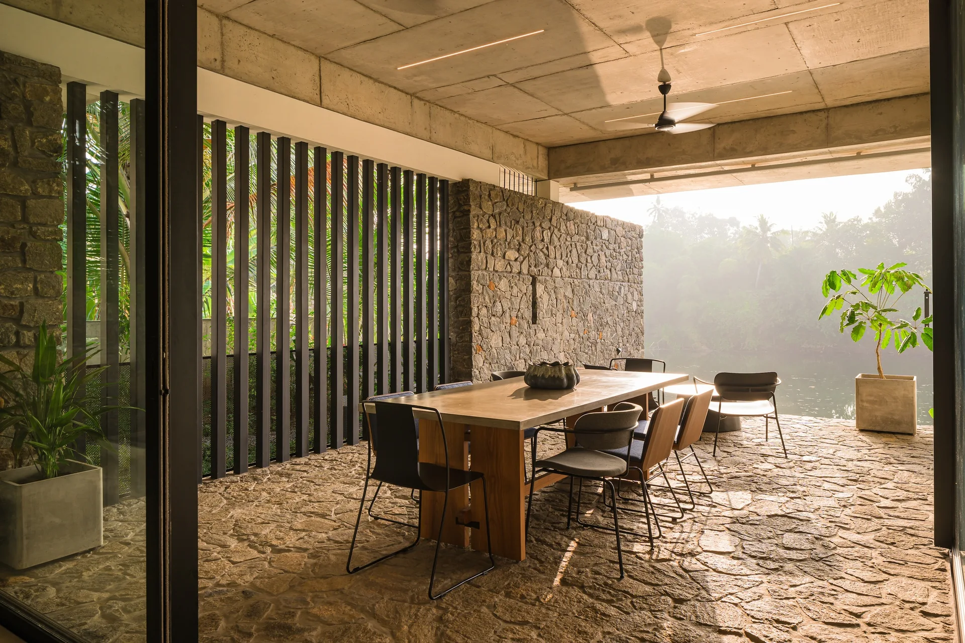 An open-air dining patio features a concrete-topped table and chairs on an irregular stone floor next to a stacked stone wall.