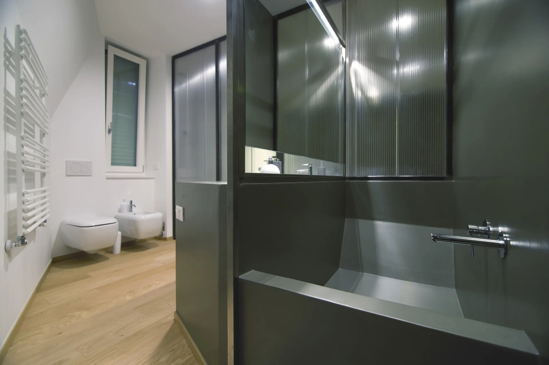A modern bathroom featuring a fluted glass partition, a dark grey built-in tub, wall-mounted white toilet fixtures, and light wood flooring.