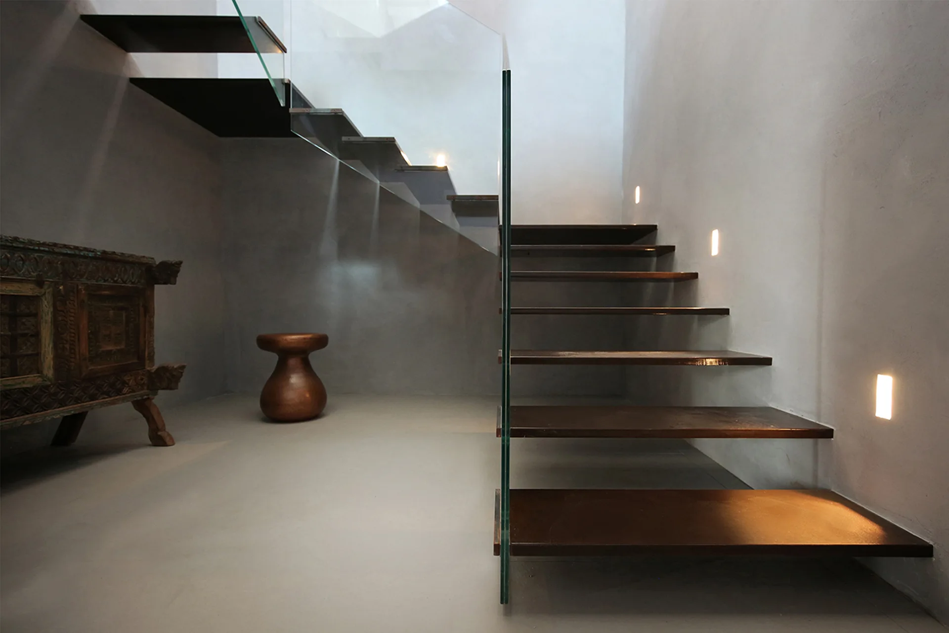 A minimalist staircase features cantilevered dark wood treads, a frameless glass railing, and warm recessed wall lights against smooth concrete-like walls.