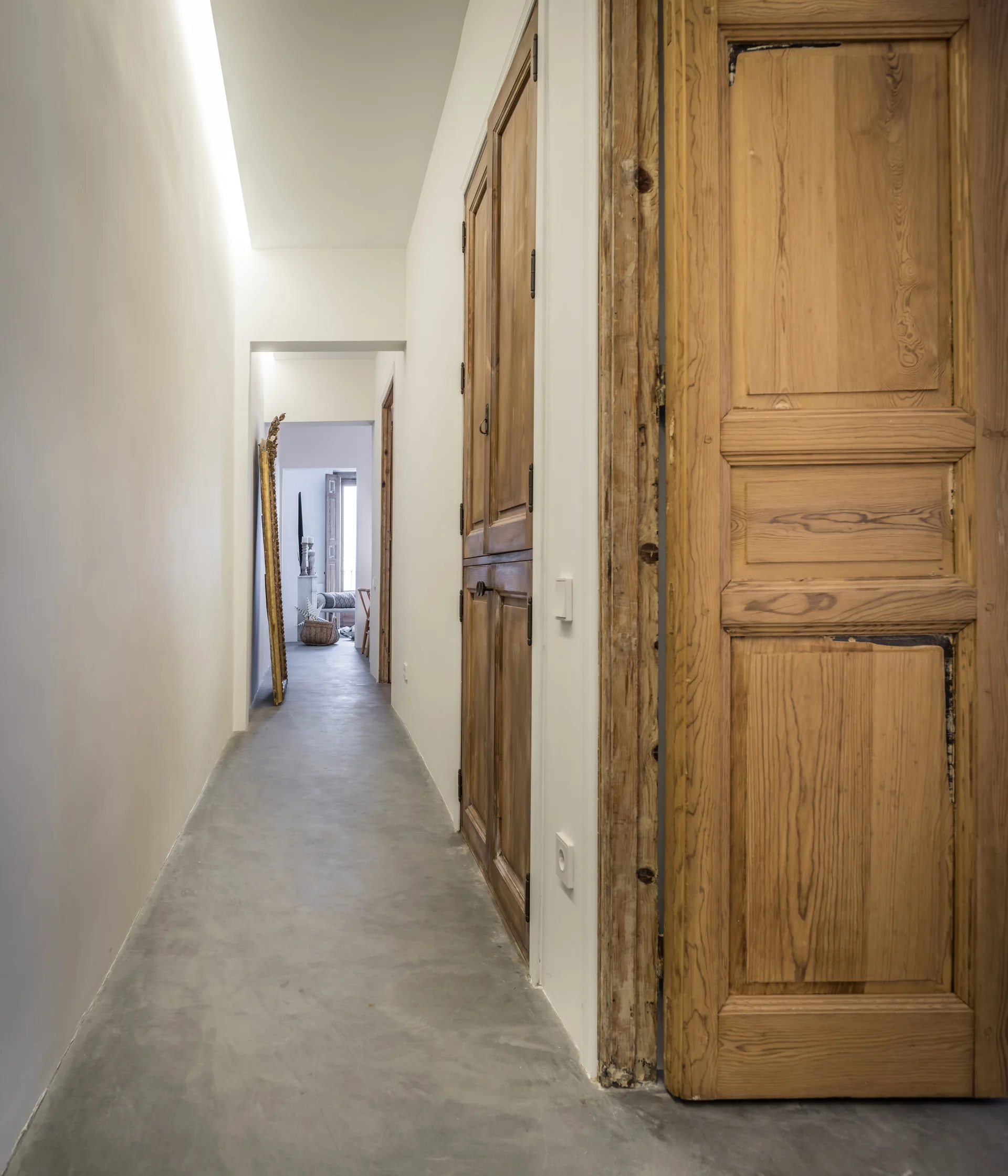 A long minimalist hallway features traditional paneled wood doors, smooth grey concrete flooring, and plain white walls with built-in cove lighting.