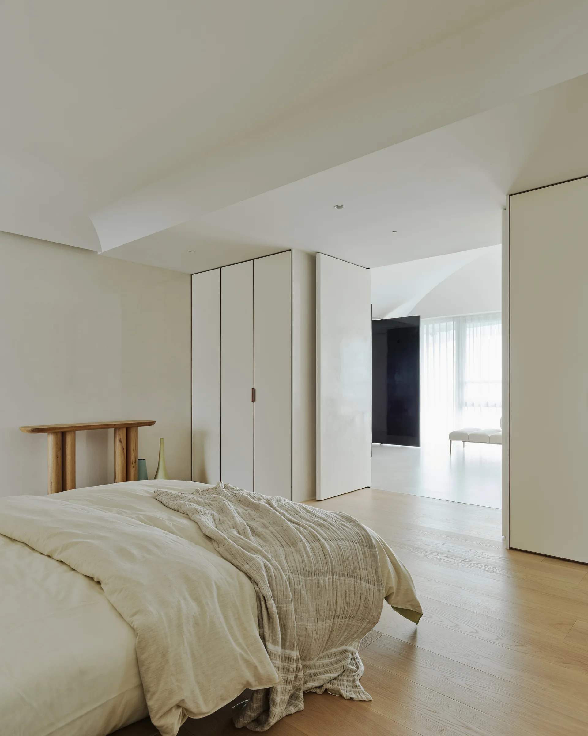 A minimalist bedroom featuring integrated off-white closets, a bed with cream linens, light wood floors, and a view into an adjacent sunlit room.