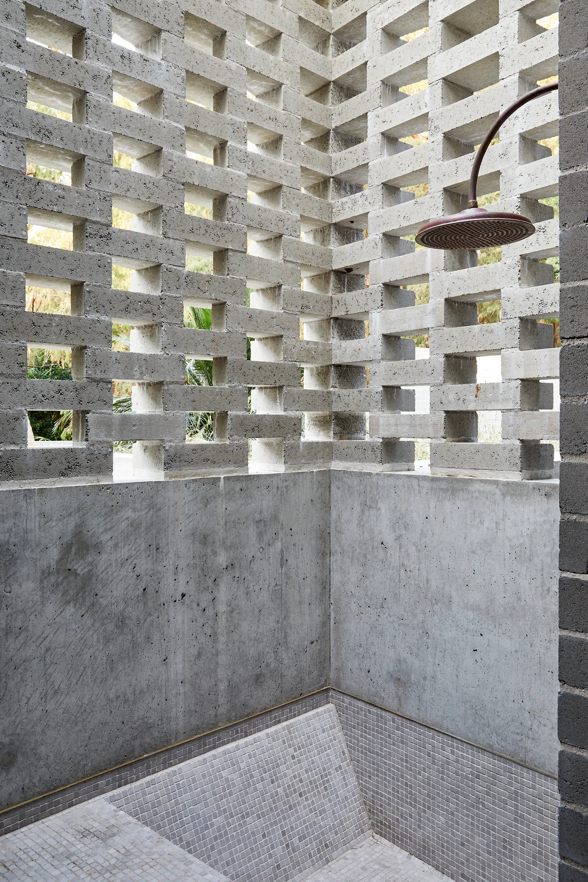 An outdoor shower features a smooth grey concrete wall topped with geometric breeze blocks and a small grey mosaic tile floor.