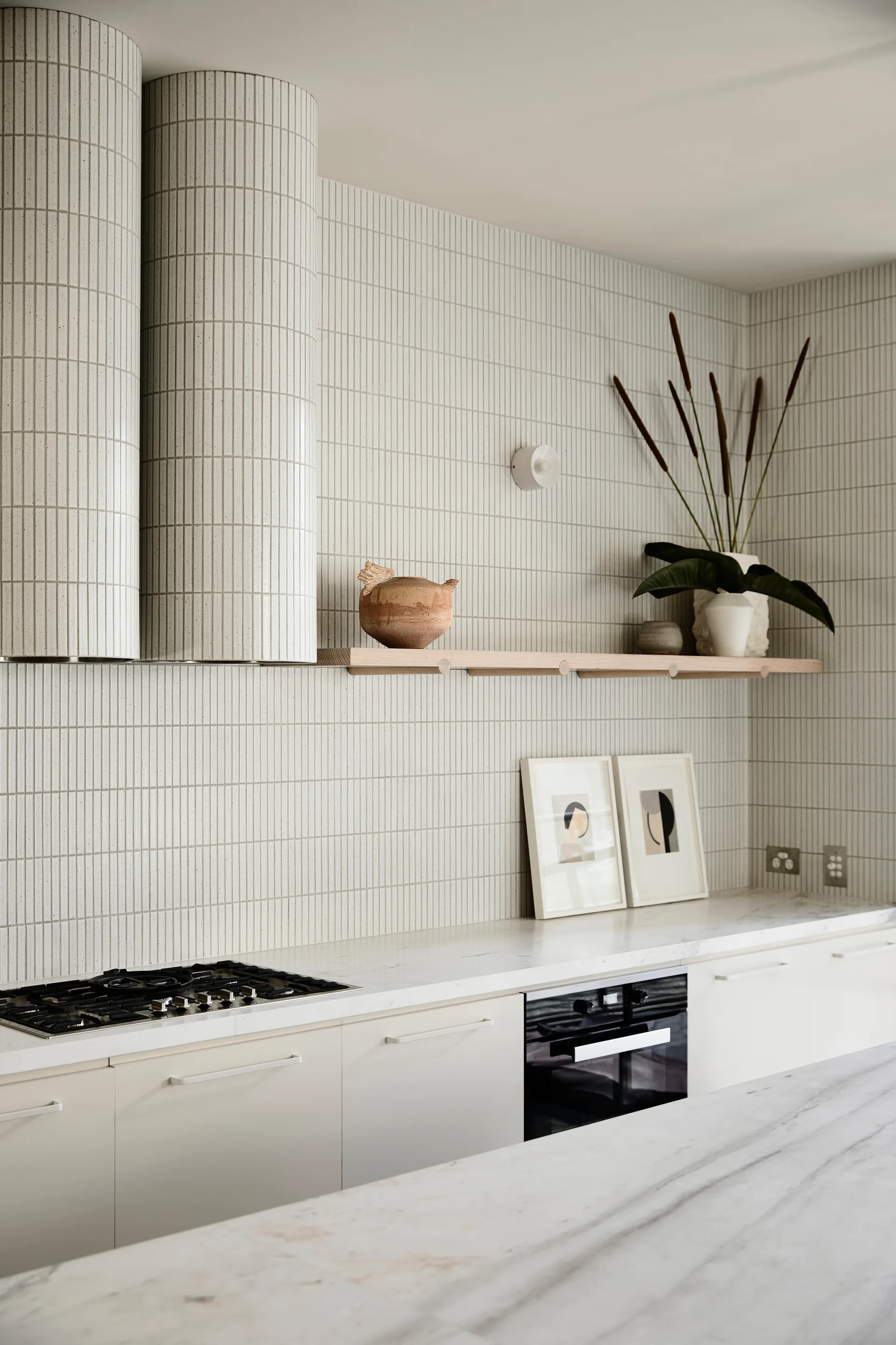 A modern kitchen featuring a vertical white tile backsplash, matching tiled range hoods, marble countertops, and a wood floating shelf.