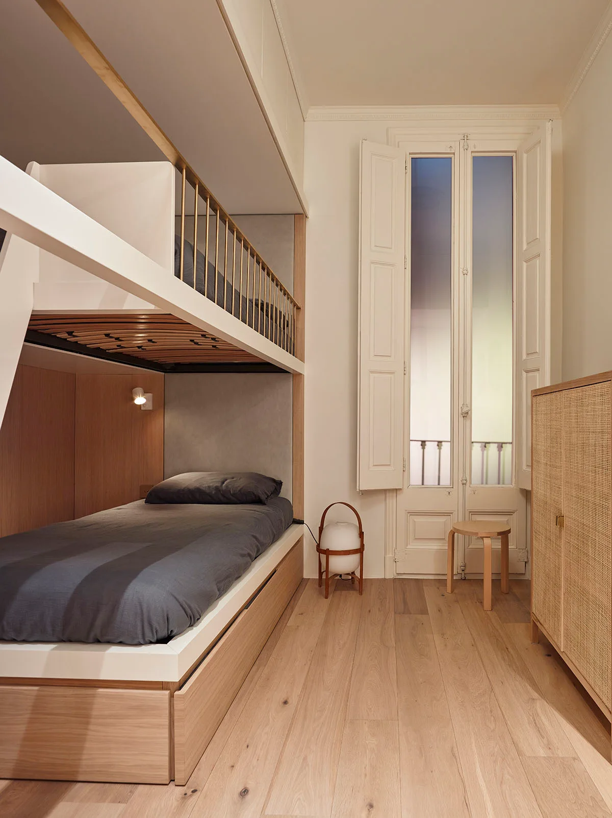 A modern bedroom features a custom light wood built-in bunk bed, wide plank flooring, a woven cane cabinet, and a tall shuttered window.