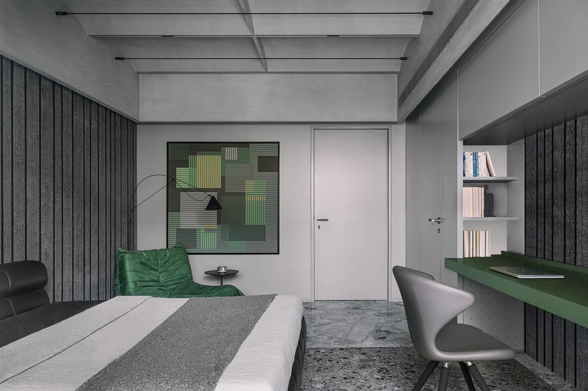 A modern grey bedroom features a built-in green desk, a matching green lounge chair, textured grey wall paneling, and an abstract artwork.