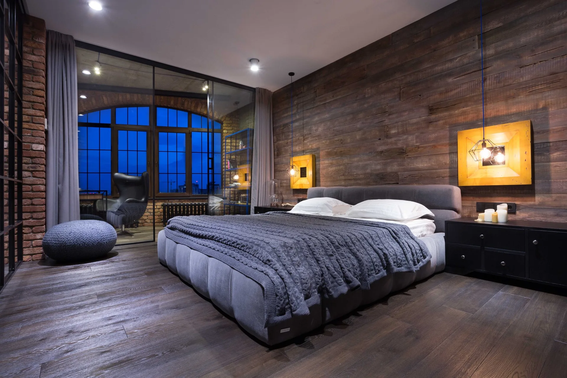 An industrial loft bedroom features a gray upholstered bed against a dark wood plank wall, black nightstands, and a floor-to-ceiling glass partition.