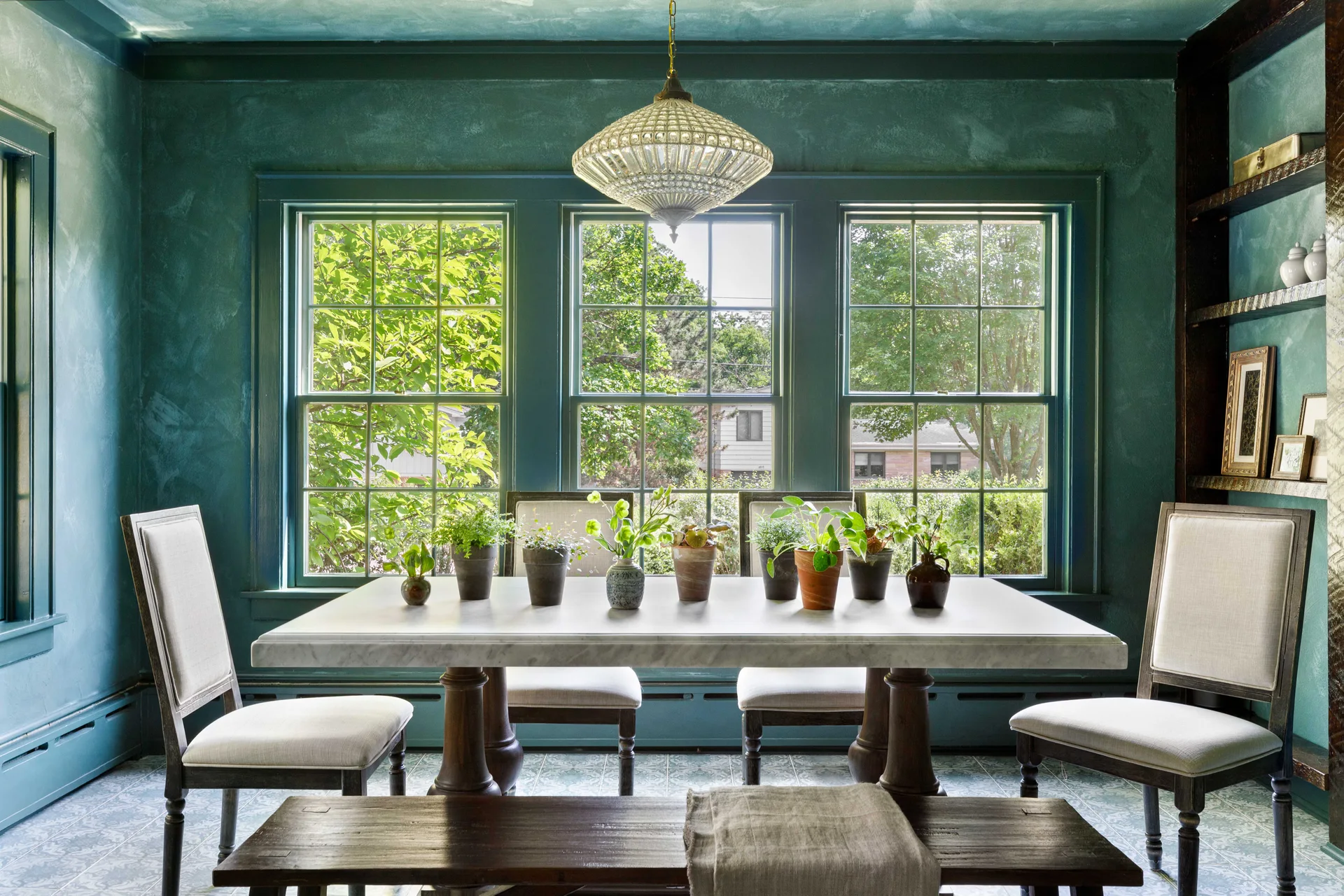 A dining room with textured teal walls and ceiling, featuring a marble table, white upholstered chairs, and a triple window overlooking trees.