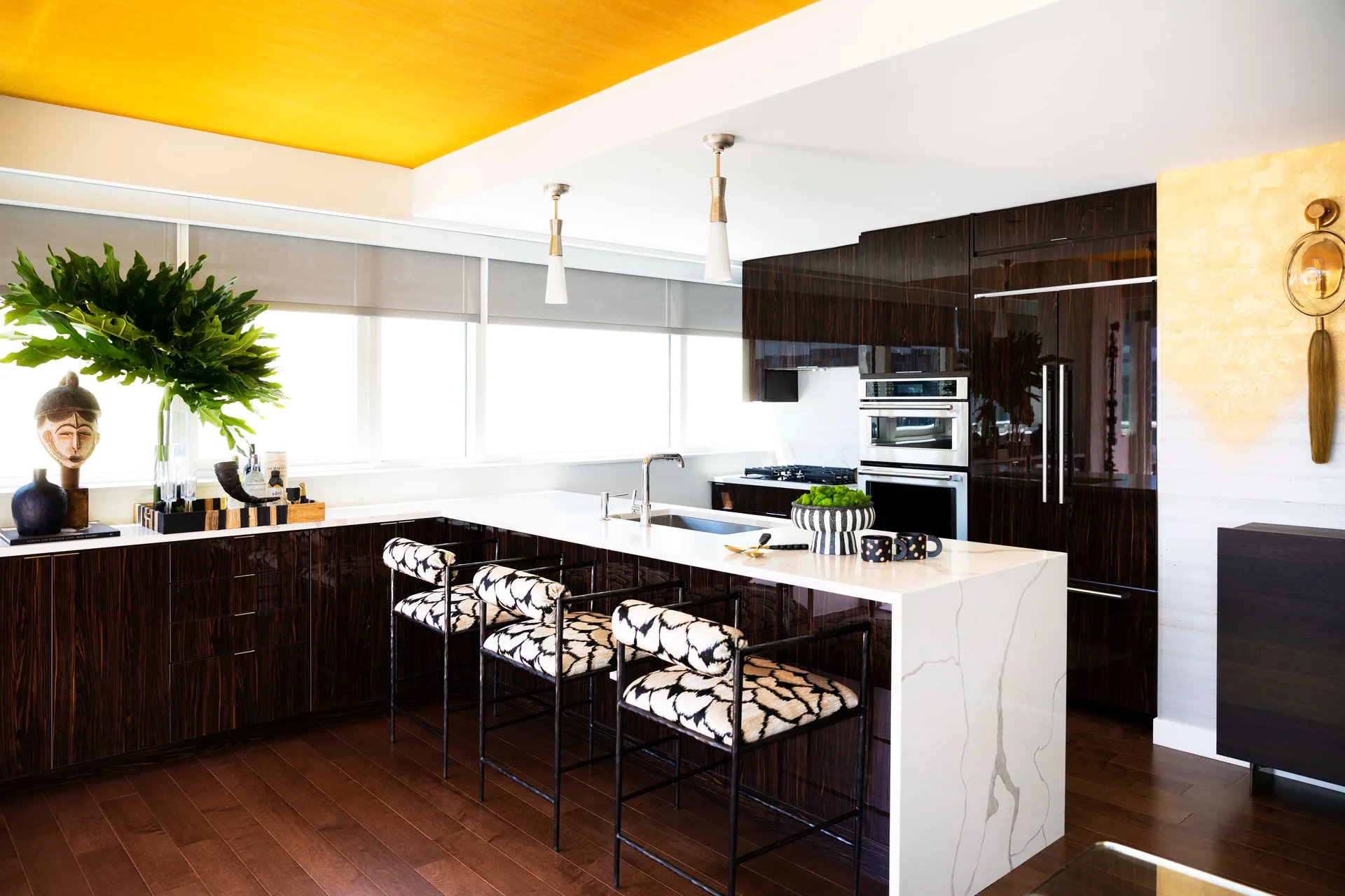 A modern kitchen features a white waterfall marble island, patterned bar stools, high-gloss dark wood cabinets, and a bright yellow ceiling.