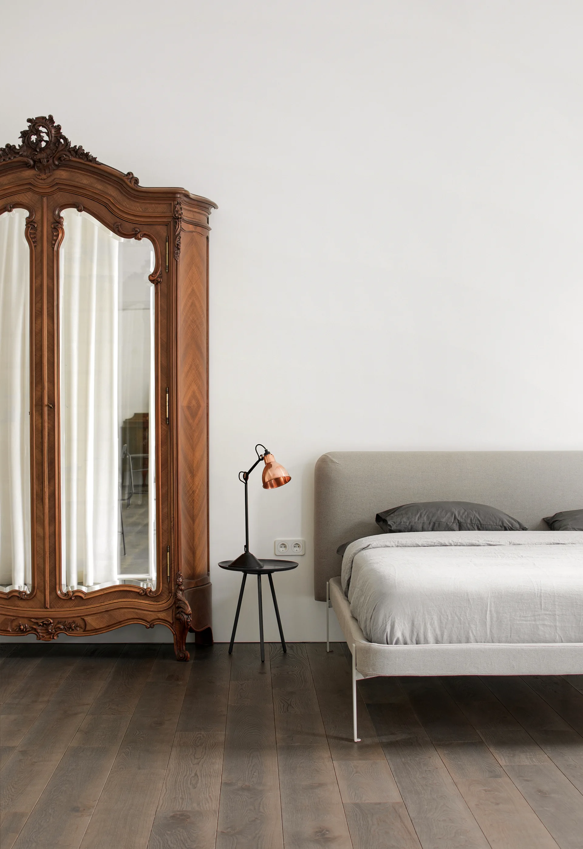 A minimalist bedroom features an ornate dark wood antique armoire next to a modern grey upholstered bed and a black tripod side table with a copper lamp.