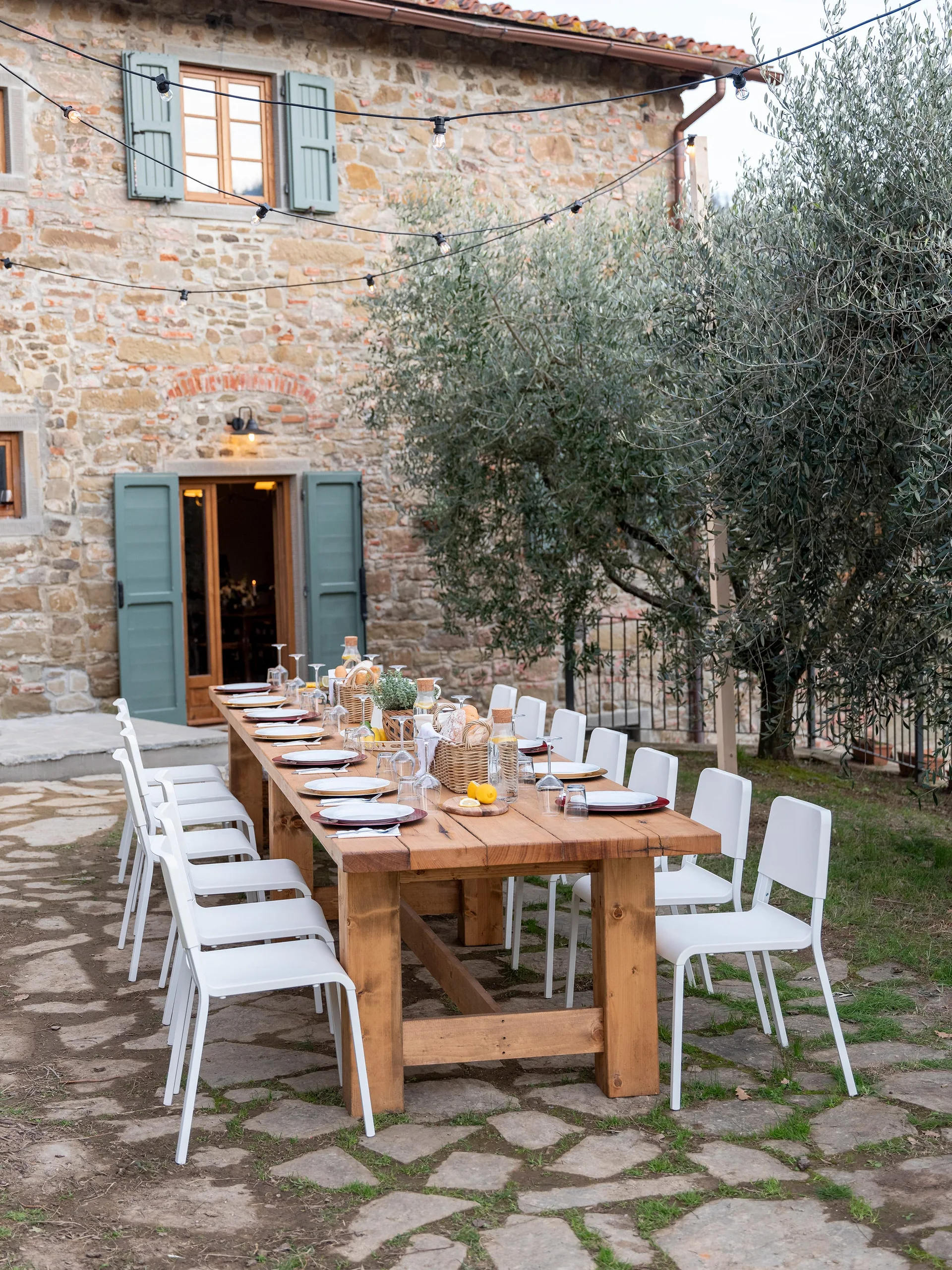 A long wooden dining table set with white chairs on a flagstone patio in front of a rustic stone building with green shutters.