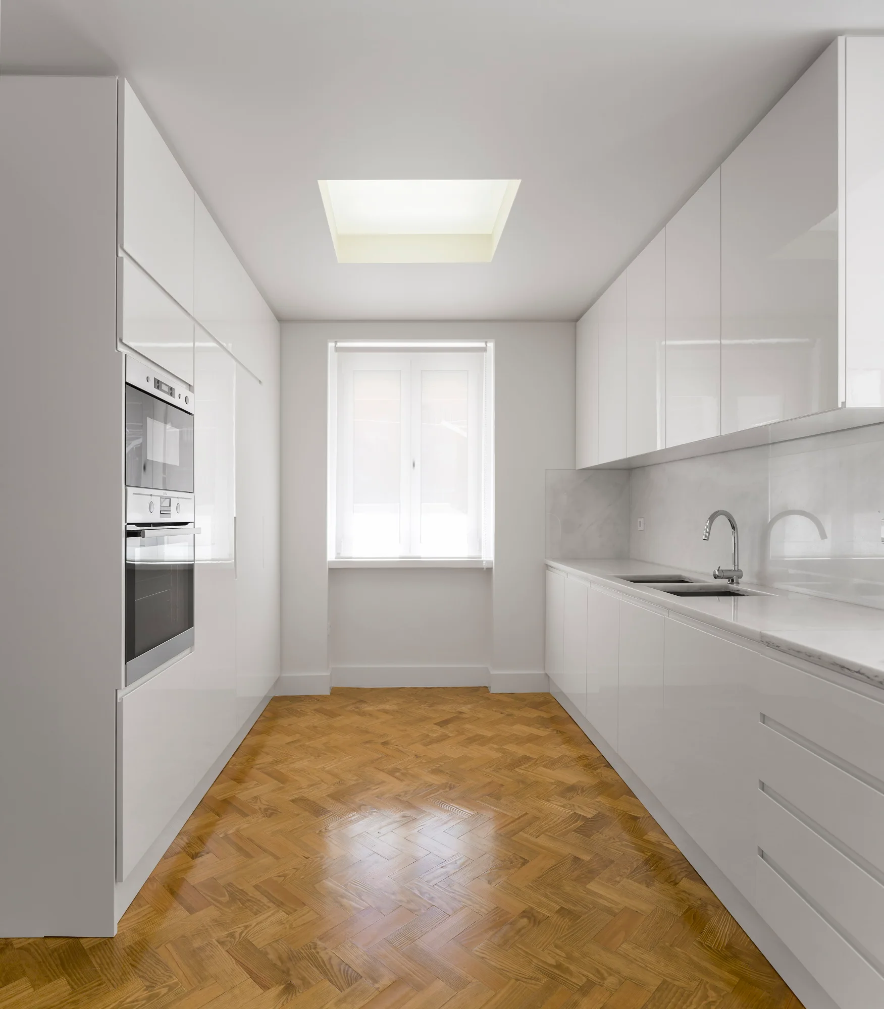 A minimalist galley kitchen featuring high-gloss white cabinets, a light stone countertop, a skylight, and a herringbone parquet wooden floor.