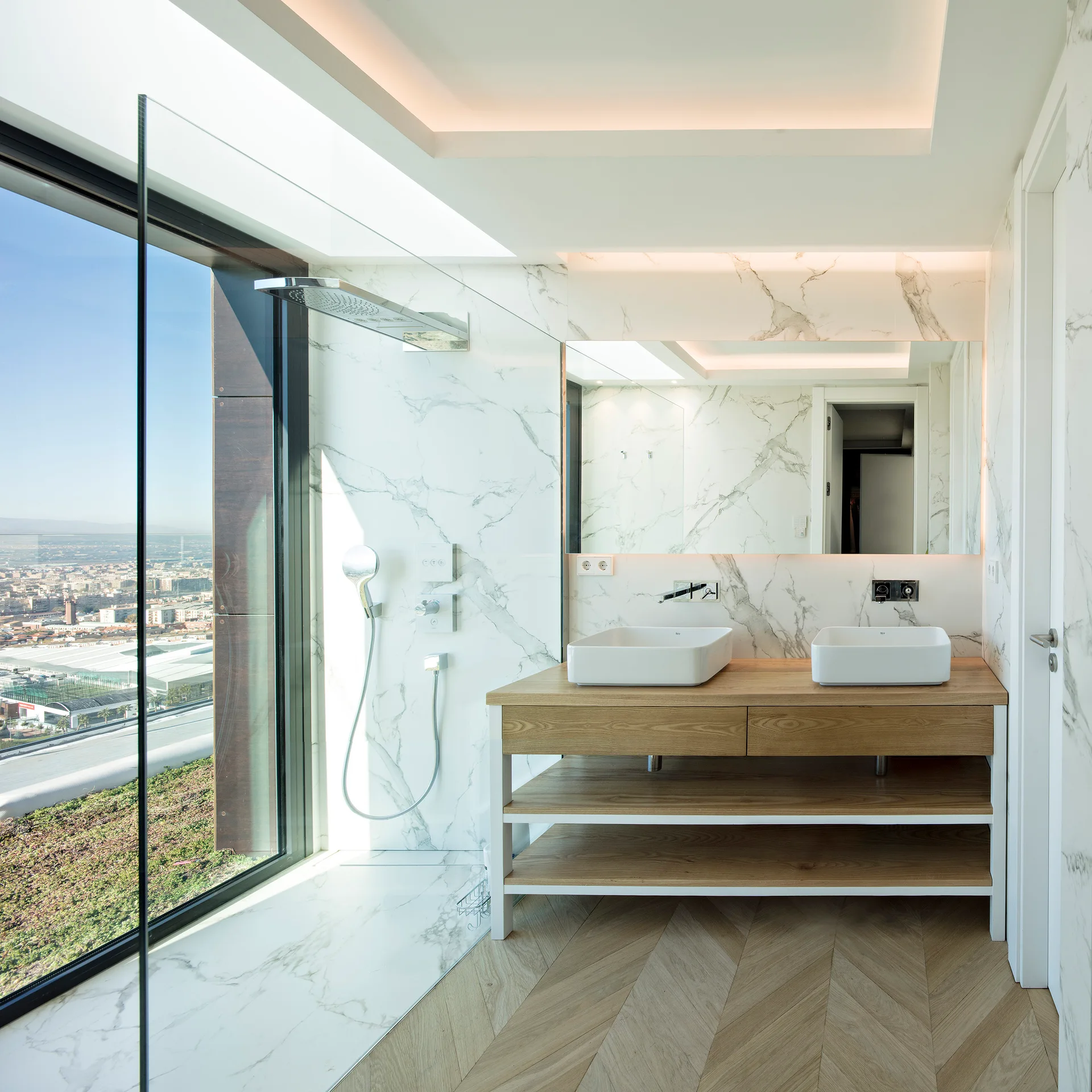 A modern bathroom features a light oak double vanity, white veined marble walls, and a glass-enclosed shower next to a large window.