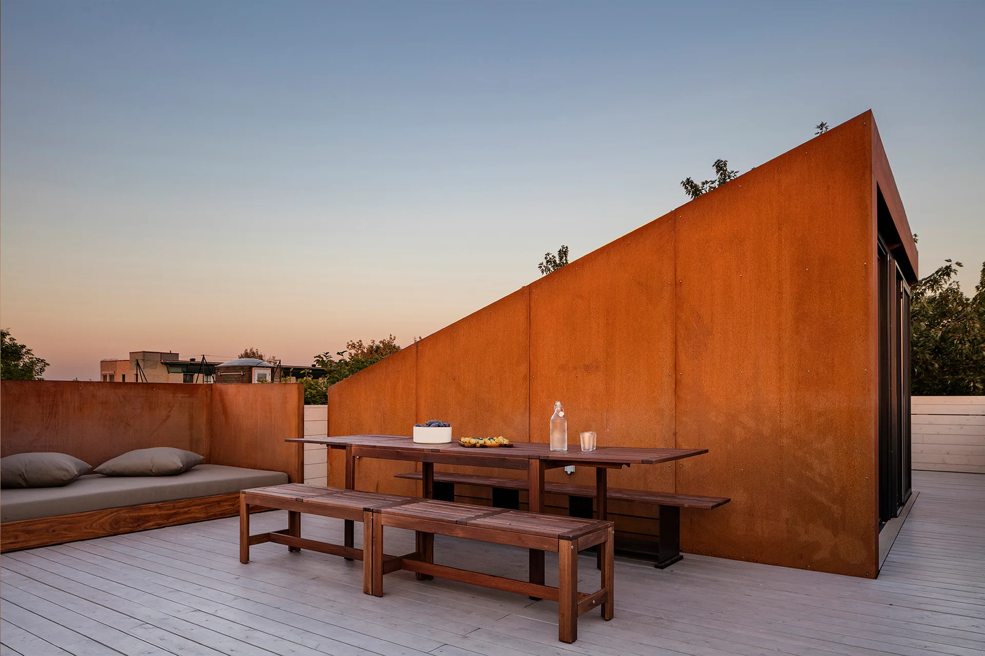 A rooftop terrace featuring a large sloped weathered steel wall, a wooden dining table with benches, and a built-in daybed on light grey wood decking.