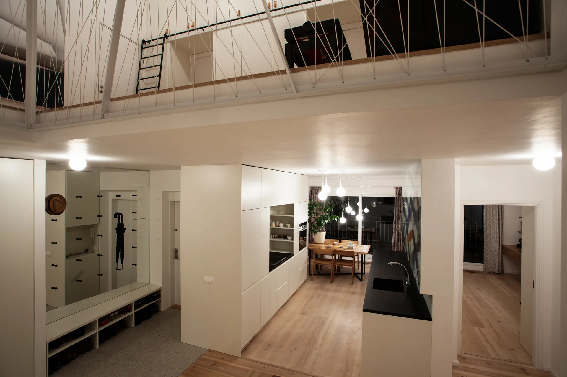 A modern white kitchen features a black island and wooden dining table, with a mezzanine loft and cable railing overhead.
