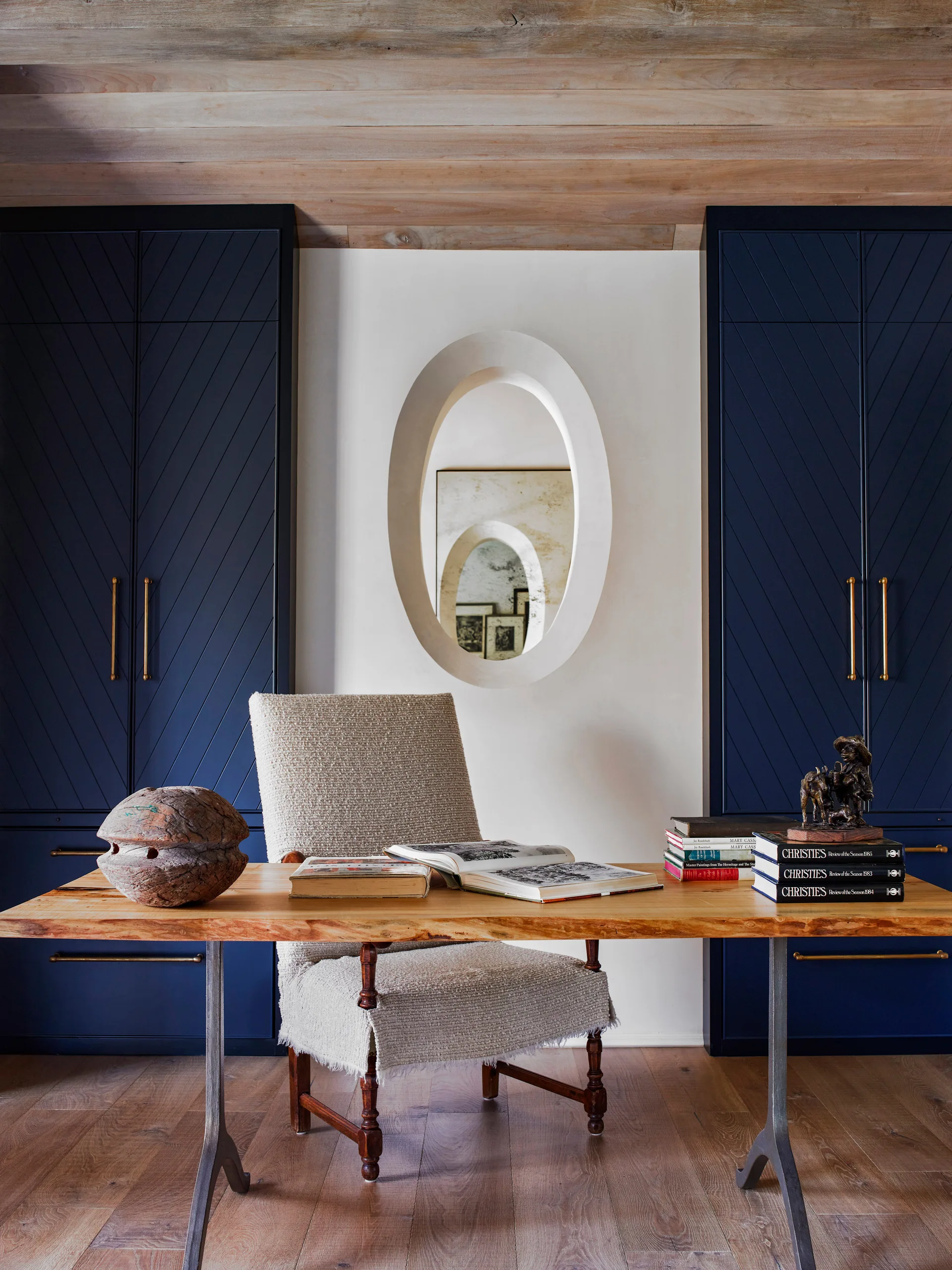 A home office featuring a live-edge wooden desk and an upholstered armchair set against dark blue cabinets and an oval wall niche.