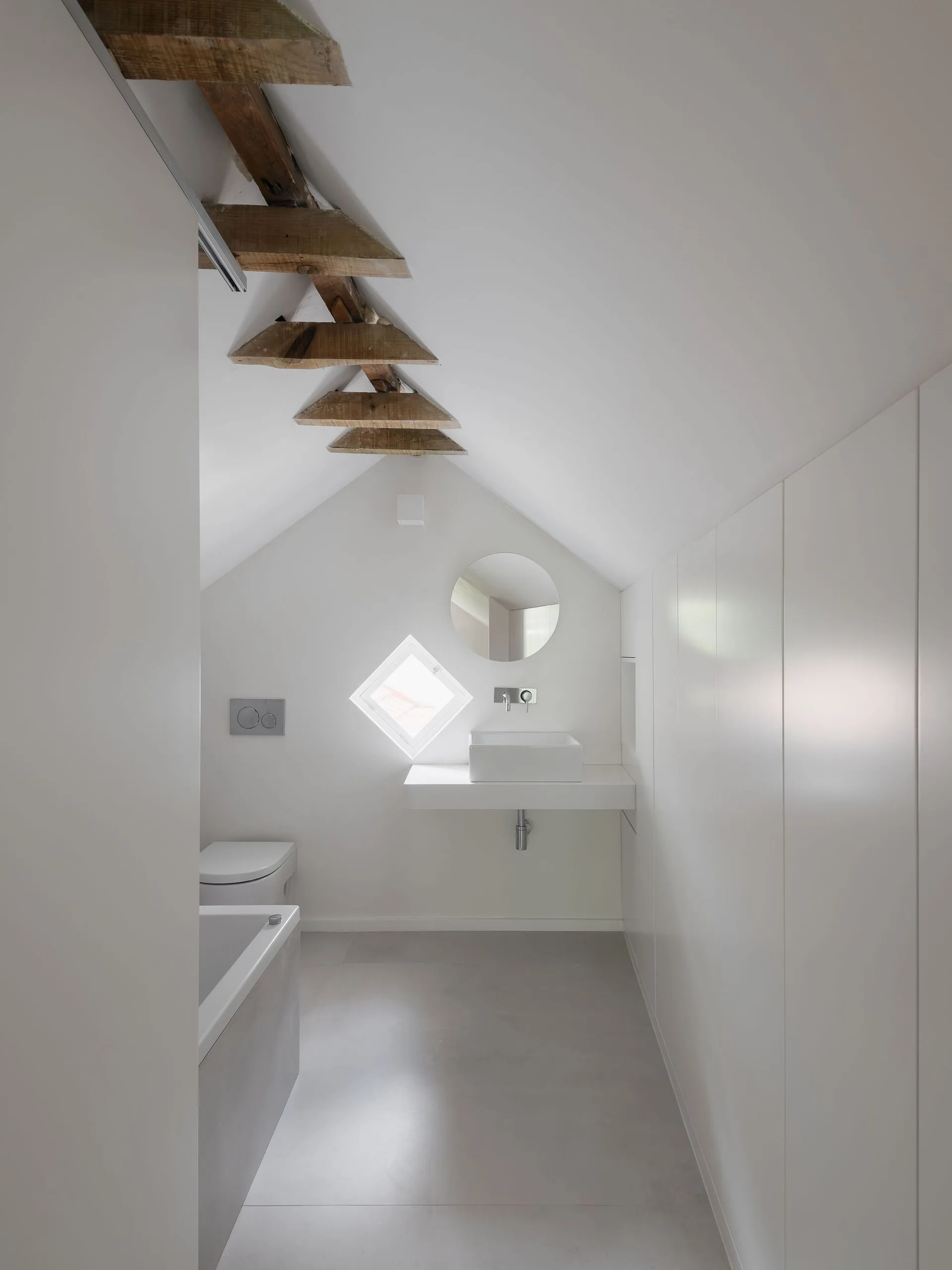 A minimalist white attic bathroom featuring a sloped ceiling with exposed wood beams, a rectangular wall-mounted sink, and a unique diamond window.