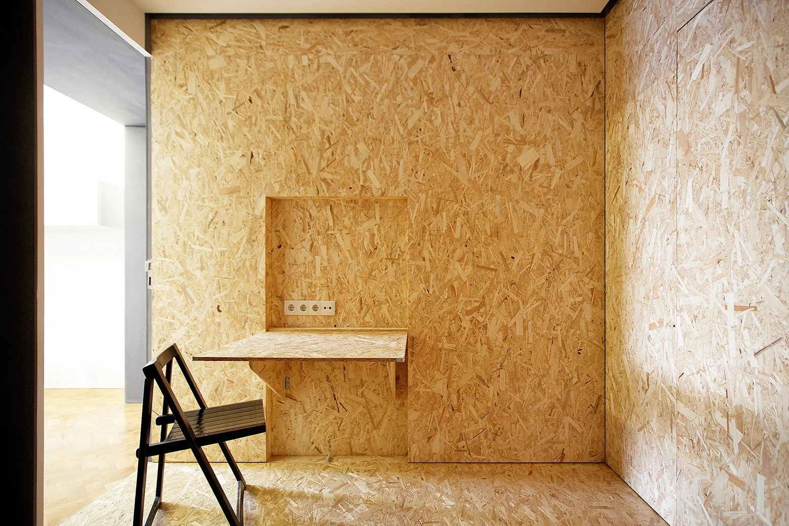 A minimalist workspace features textured OSB wood paneling on the walls and floor, a built-in niche desk, and a black folding chair.