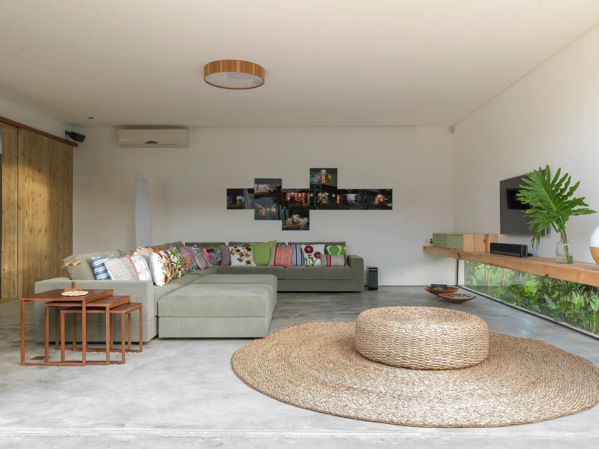 An olive green sectional sofa sits on a concrete floor next to a large circular woven rug, a wooden floating shelf, and a low horizontal window.