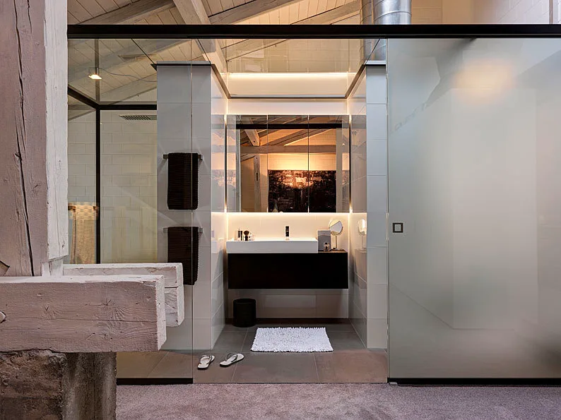 A modern bathroom features a dark floating vanity, a frosted glass sliding door, and a mirrored cabinet illuminated by integrated cove lighting.