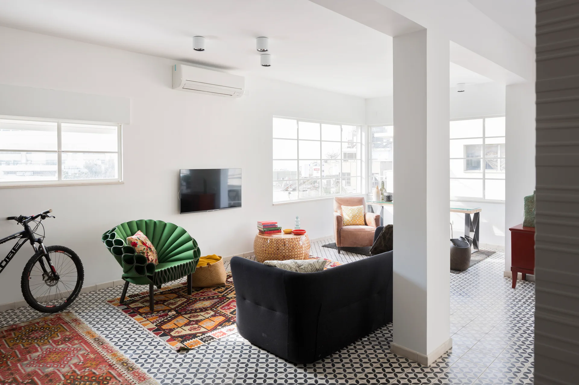 An eclectic living room features geometric tile flooring, a dark sofa, a fan-shaped green armchair, and layered patterned rugs under large windows.