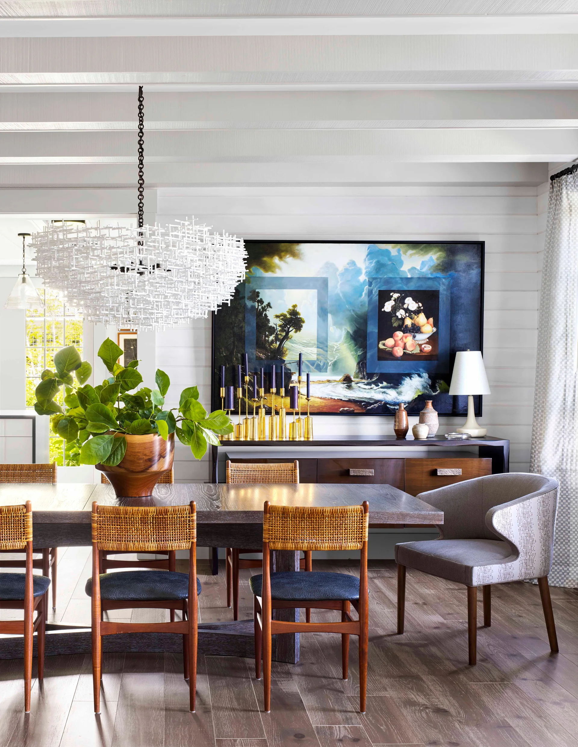A dining room features a dark wood table with cane-backed chairs, a large white sculptural pendant light, and a horizontal landscape painting.