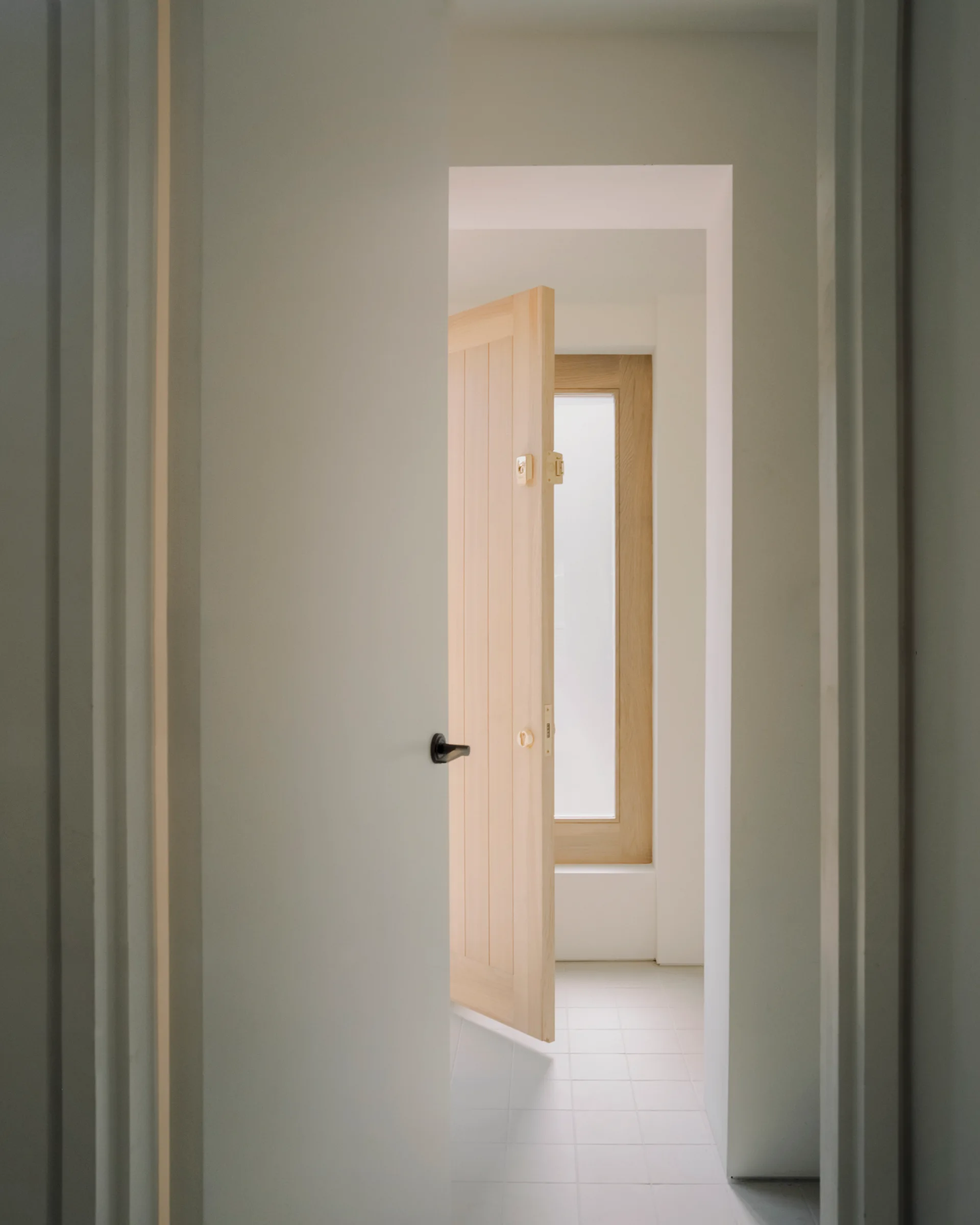 A view through a white doorway looking toward an open light wood door and a wood-framed window over a white square tiled floor.