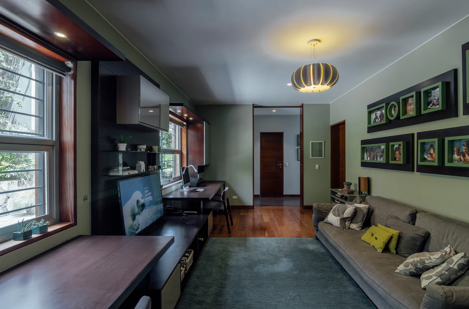 A transitional home office and living space features sage green walls, a long built-in dark wood desk, and a gray sofa.