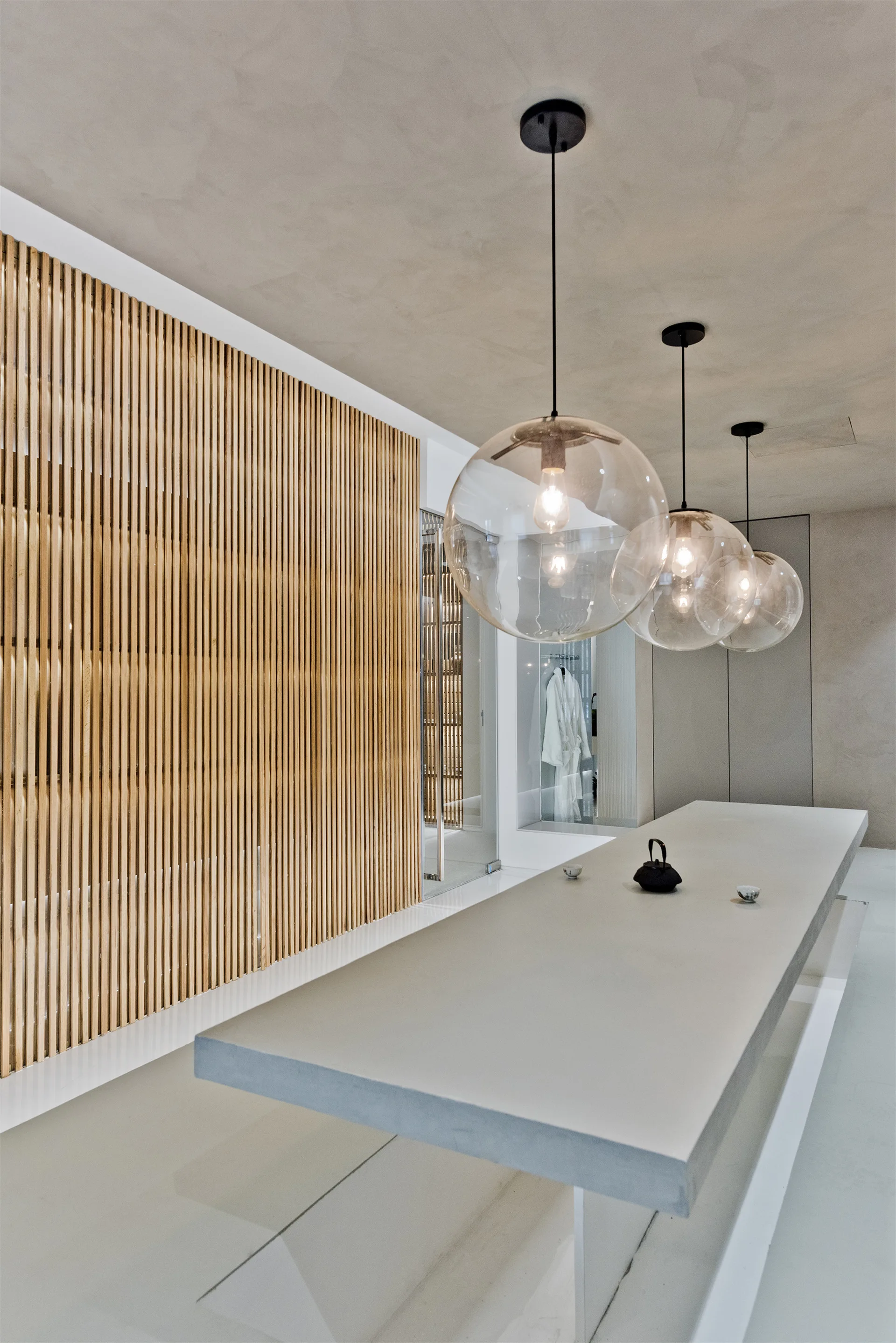 A minimalist dining area featuring a long grey table, a vertical wood slat wall, and three clear glass globe pendant lights.
