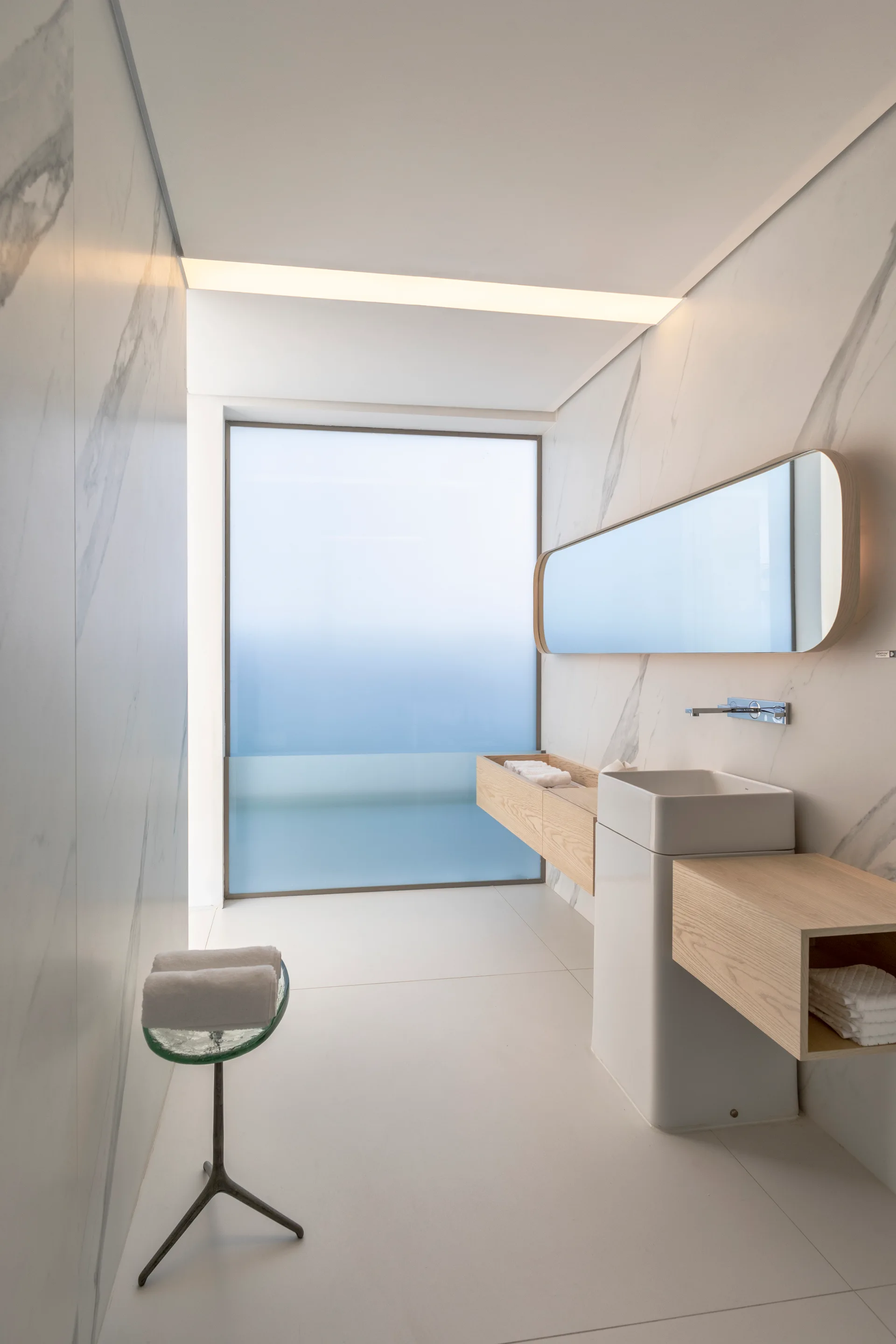 A minimalist bathroom featuring marble-veined wall tiles, a white pedestal sink flanked by light wood floating shelves, and a large frosted glass window.