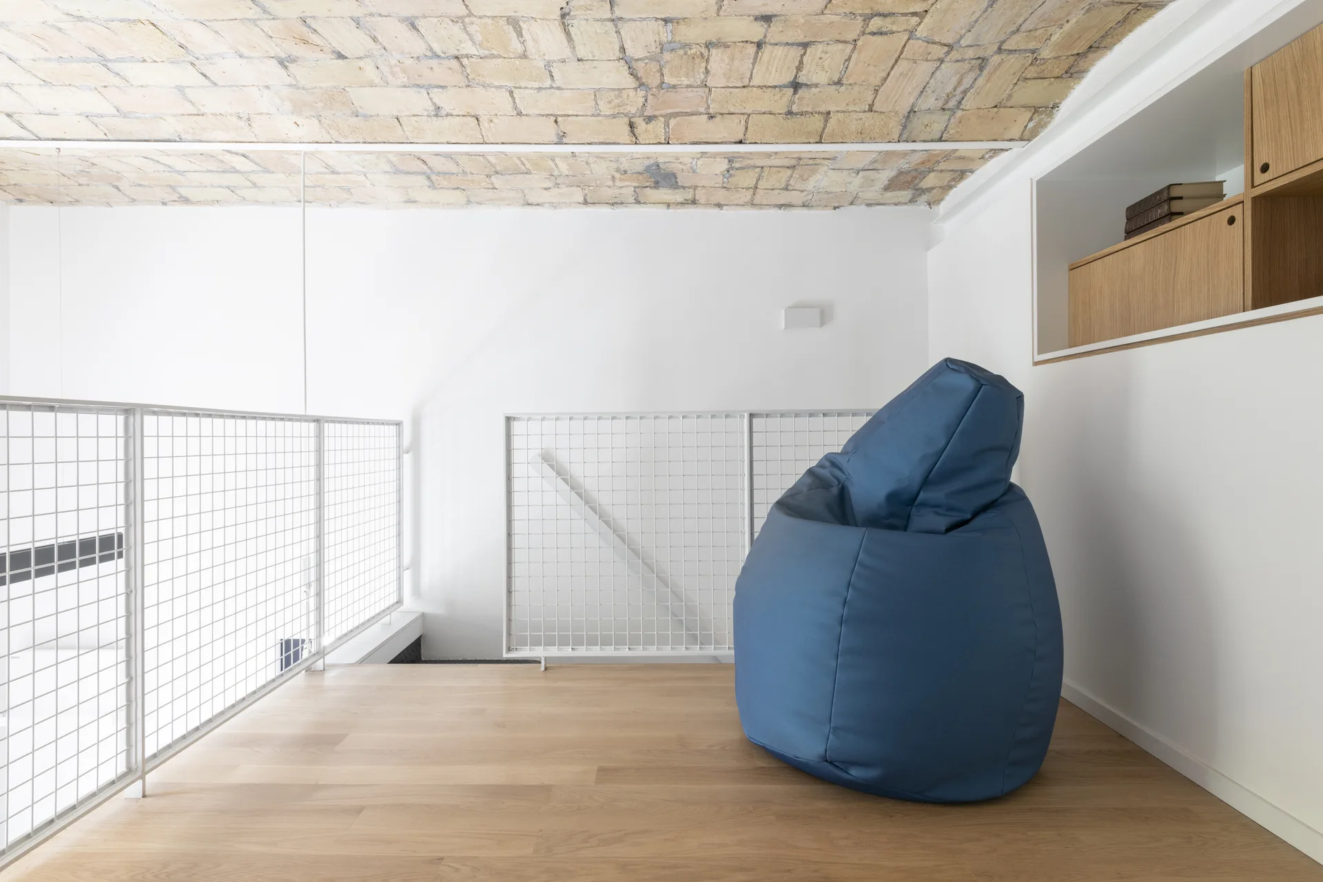A minimalist landing with a blue beanbag chair, white wire mesh railing, wooden floors, and an exposed brick vaulted ceiling.