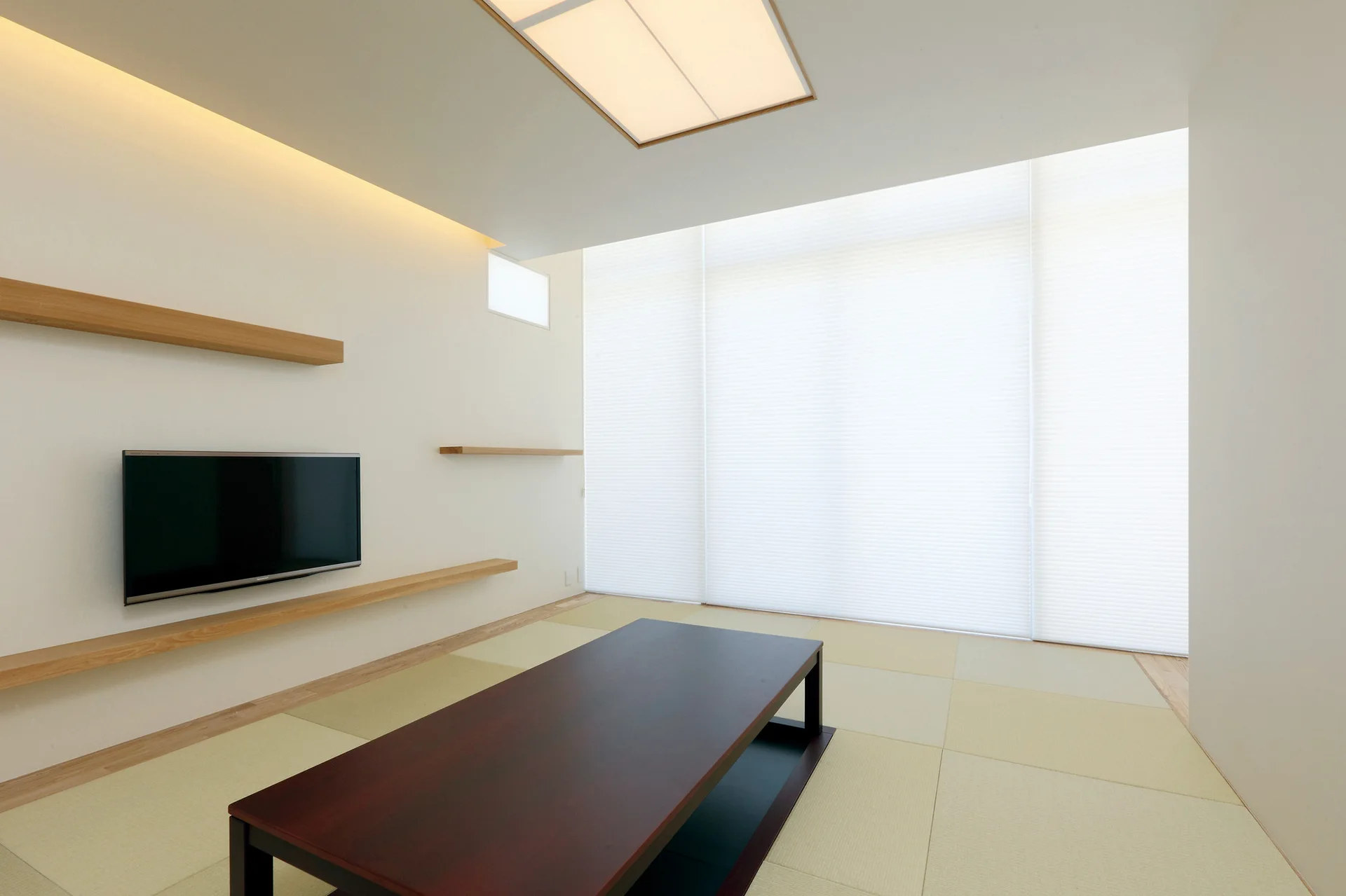 A minimalist room features tatami mat flooring, a low dark wood coffee table, floating wall shelves, and a wall-mounted television.