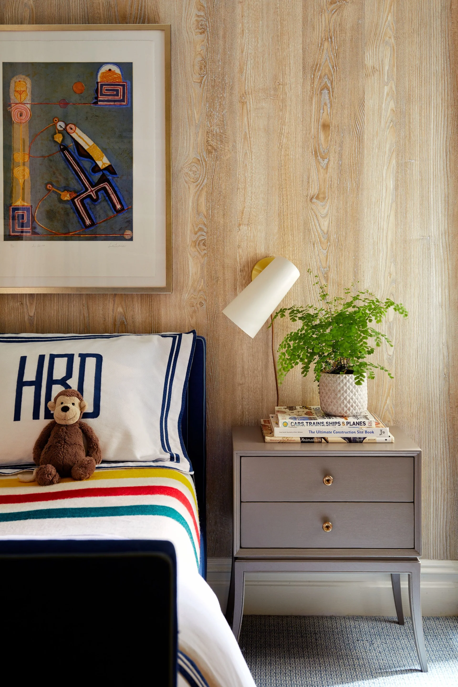 A children's bedroom corner featuring a wood-paneled wall, a grey nightstand with books and a plant, and a striped bed.