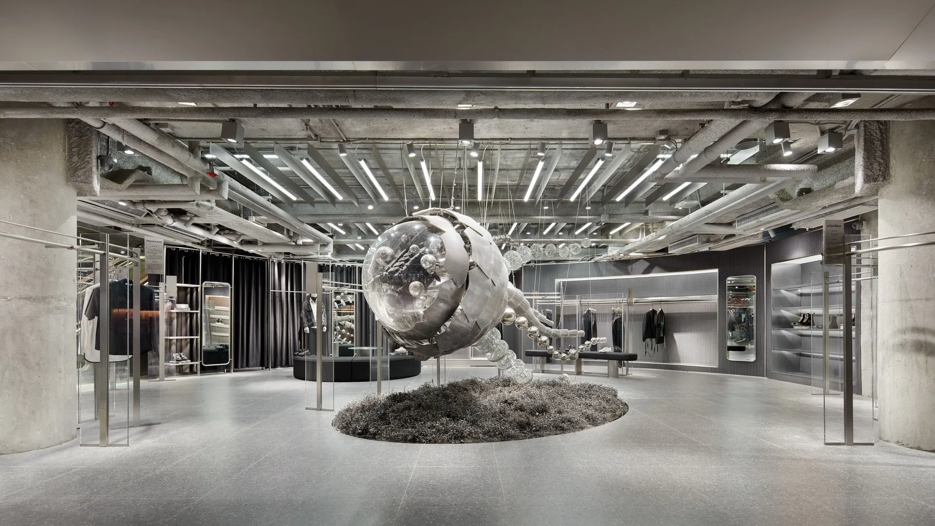 An industrial retail showroom features a large central metallic sculpture over metal shavings beneath an exposed concrete ceiling with linear lighting.