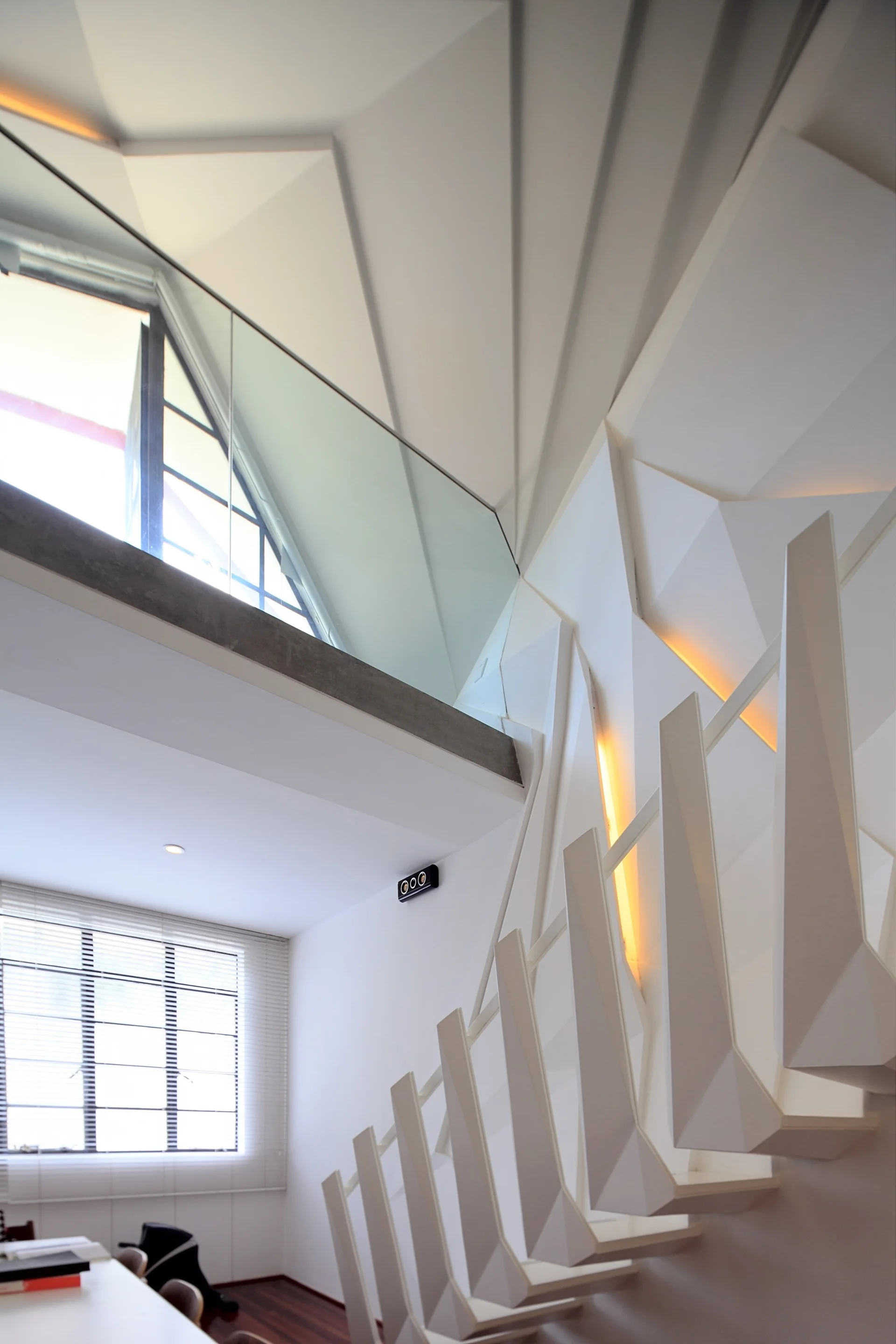 A minimalist white staircase features faceted wall paneling with integrated warm yellow lighting, an angular railing, and a glass partition.