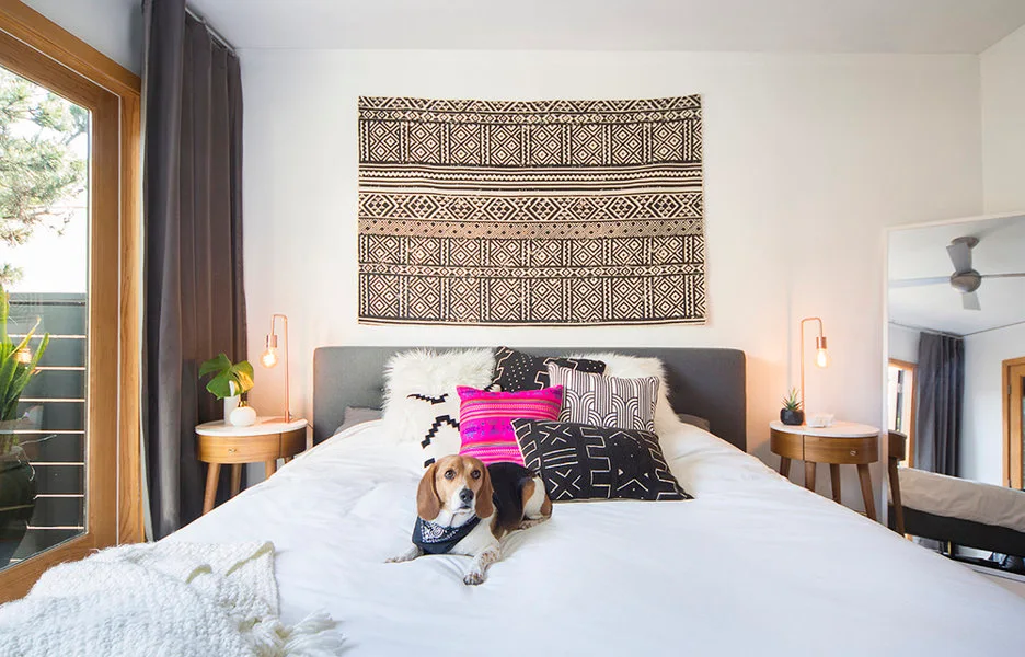 An eclectic bedroom features a bed with patterned throw pillows, a graphic black-and-white wall tapestry, and matching round wooden nightstands.