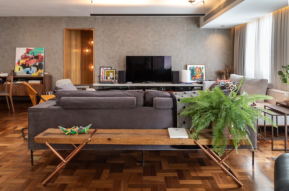 A gray living room features a textured plaster wall, a dark gray sofa, a wood parquet floor, and a wooden bench with copper legs.