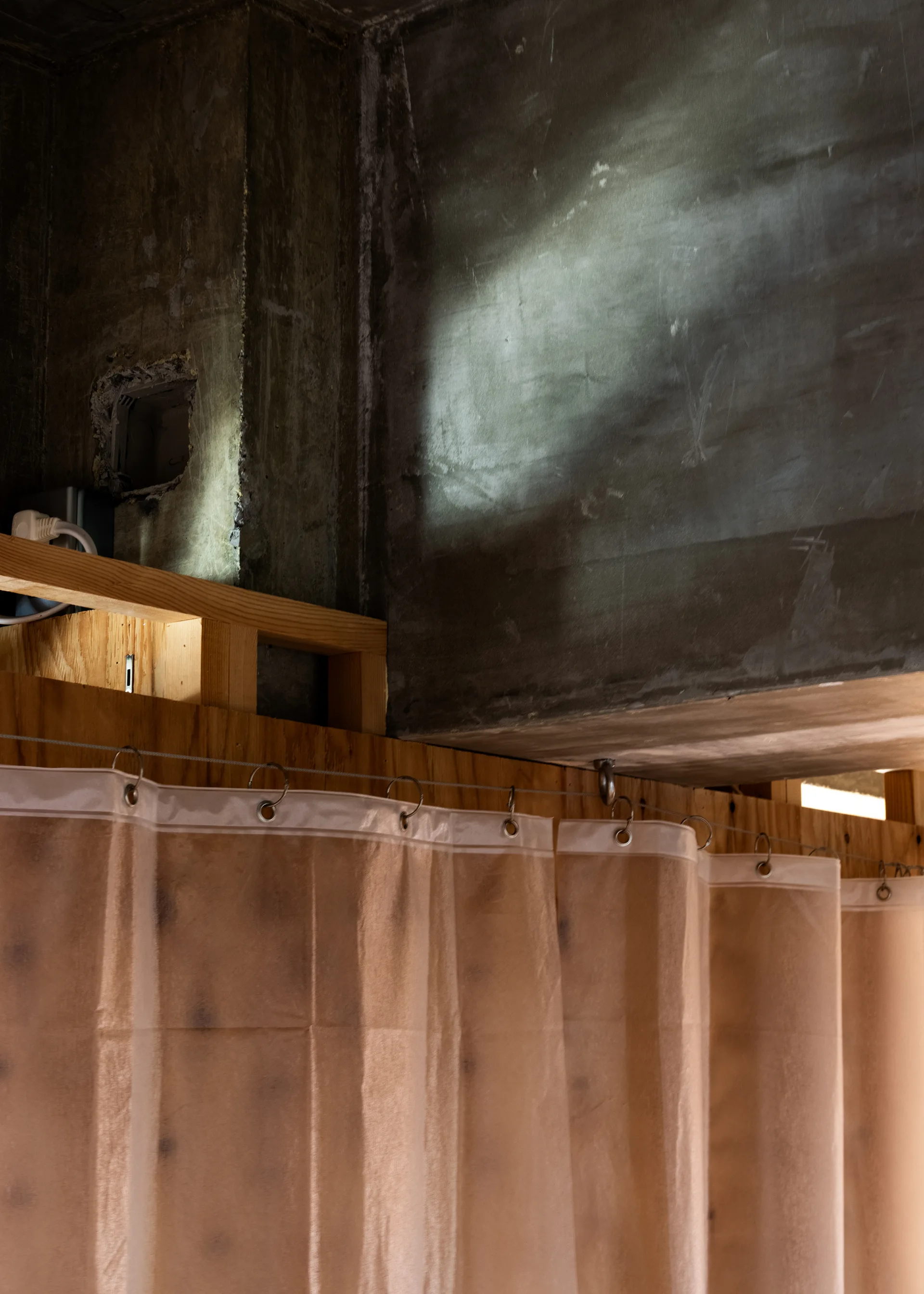 A sheer tan curtain hangs from a metal wire cable against an unfinished concrete wall and exposed wood framing.