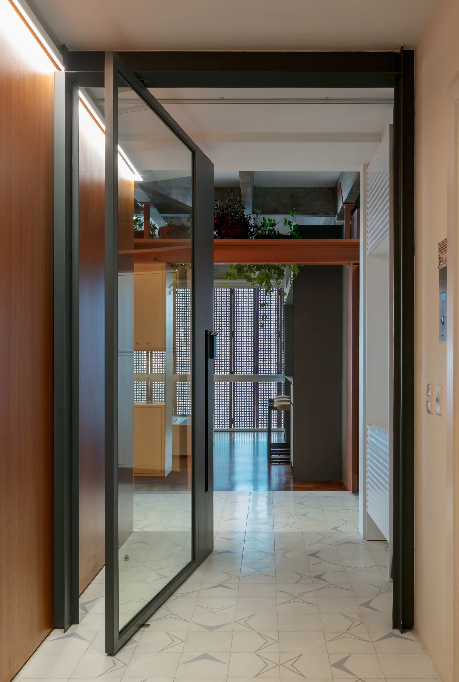 A pivoting glass door opens into an entryway with star-patterned floor tiles, wood wall panels, and an exposed concrete ceiling beam.