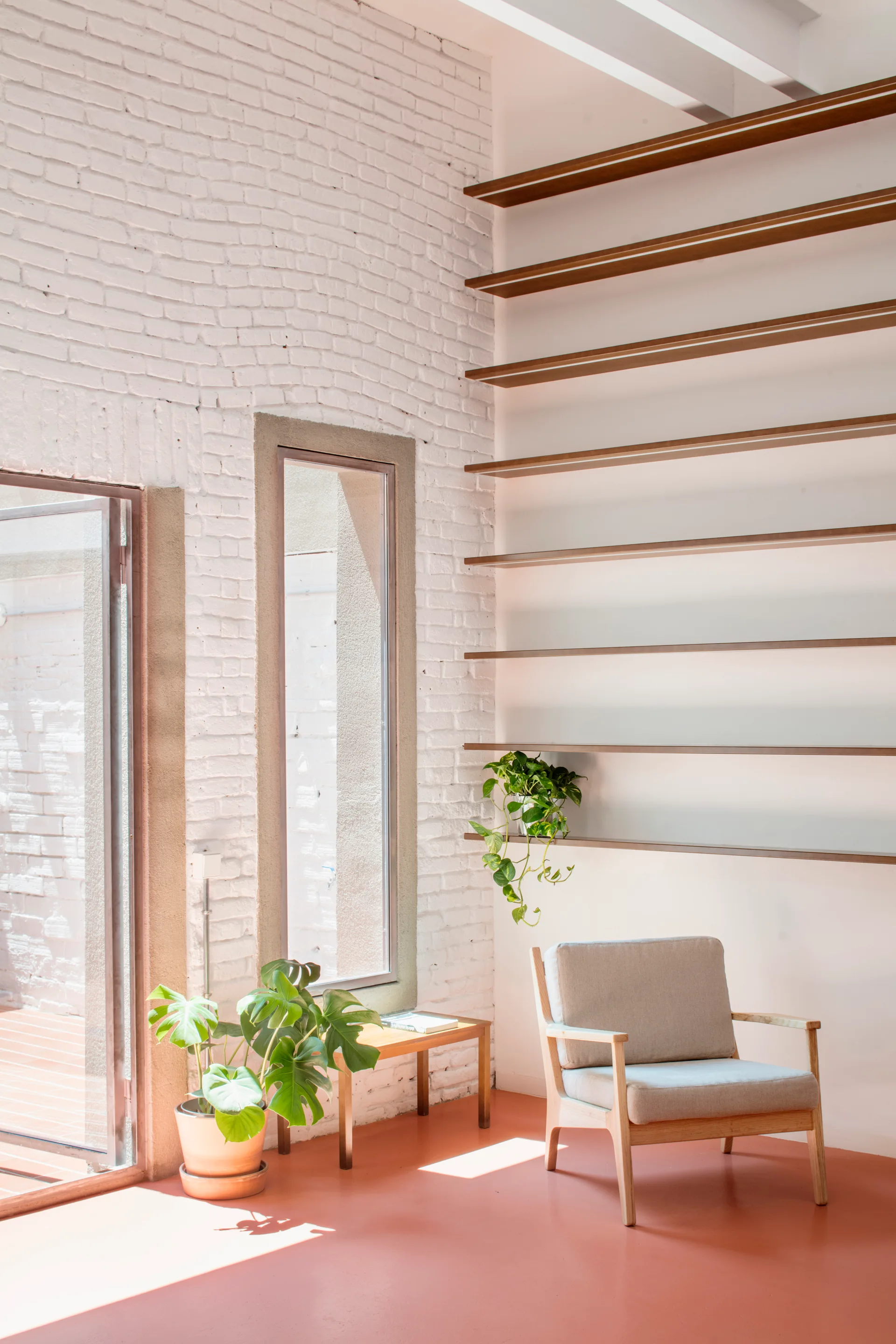 A minimalist living space features a grey armchair on a terracotta floor next to a white painted brick wall with floating wood shelves.