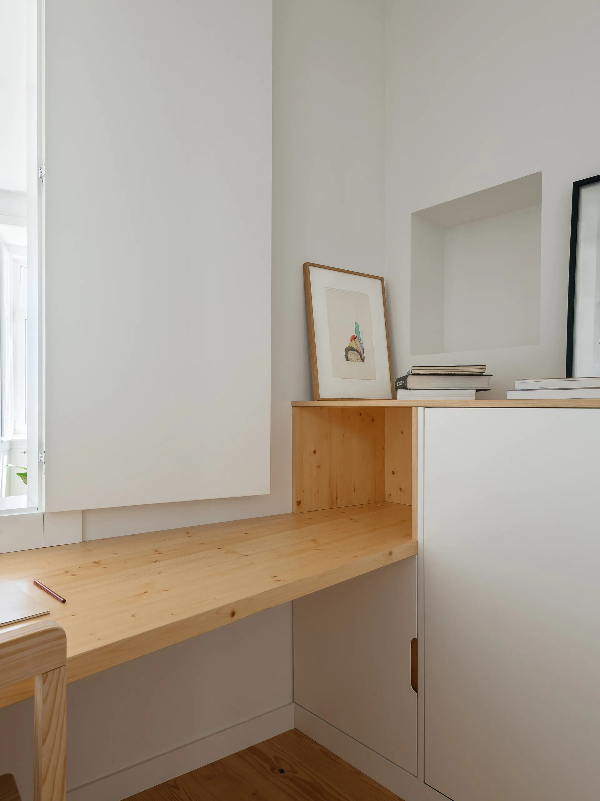 A minimalist home office workspace features a custom light wood desk built into white cabinetry beside a white wall with a small niche.