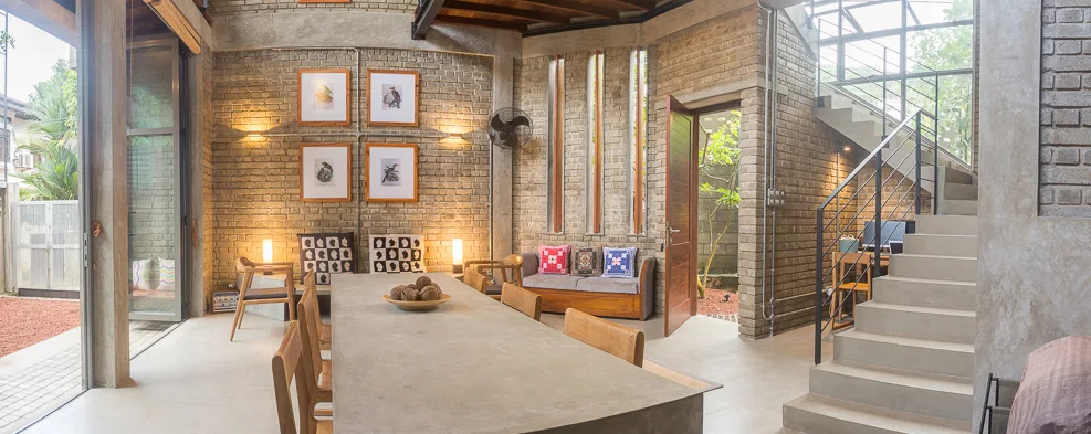 An industrial open-plan dining space featuring a long concrete table, exposed brick walls, a wooden door, and a concrete staircase.