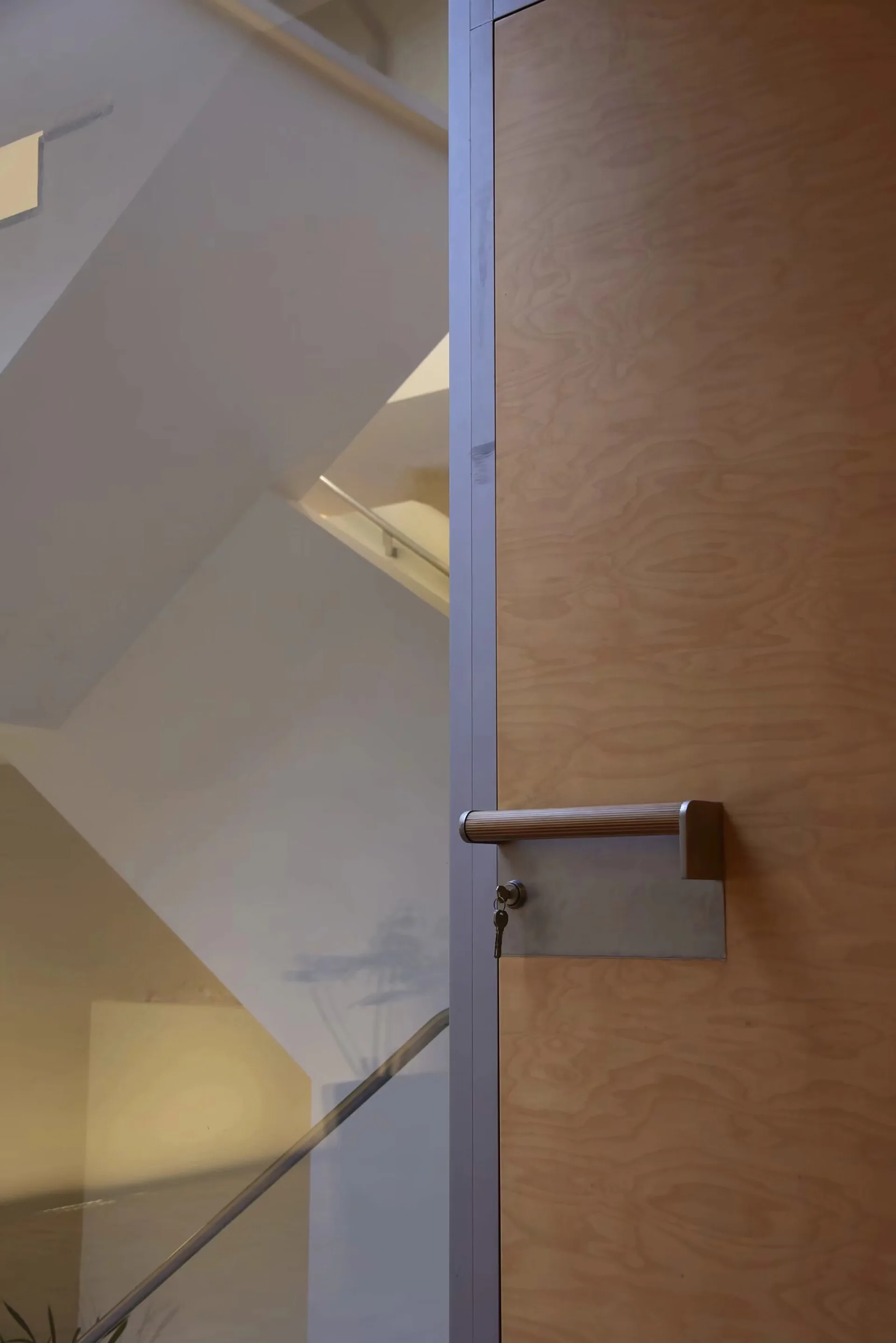 A close-up of a wood-grained door with a modern metal handle and a key, situated next to a glass partition looking onto a bright indoor staircase.