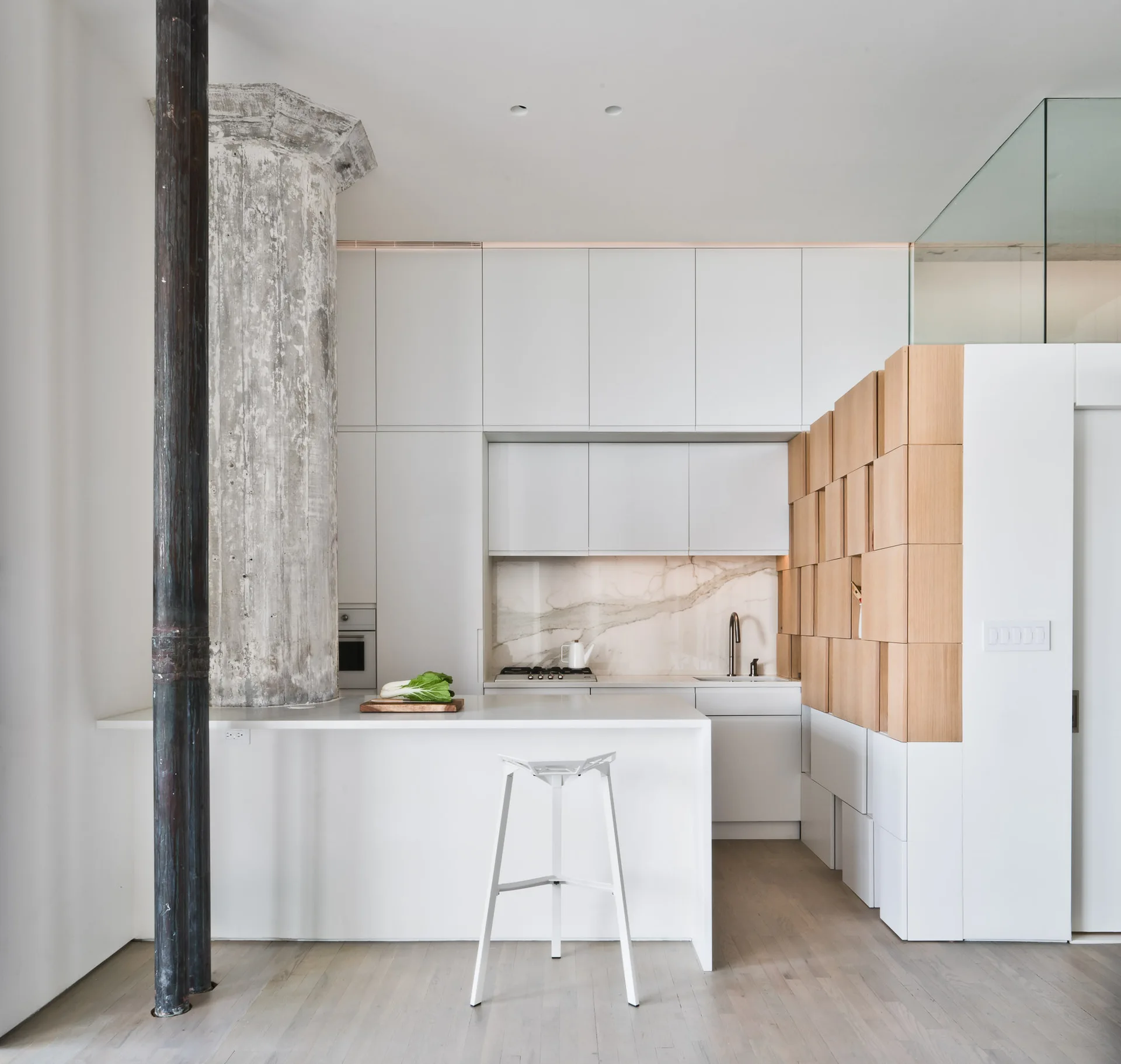 A minimalist white kitchen features a large raw concrete column, white slab cabinetry, an island with a metal stool, and a tiled wood partition.