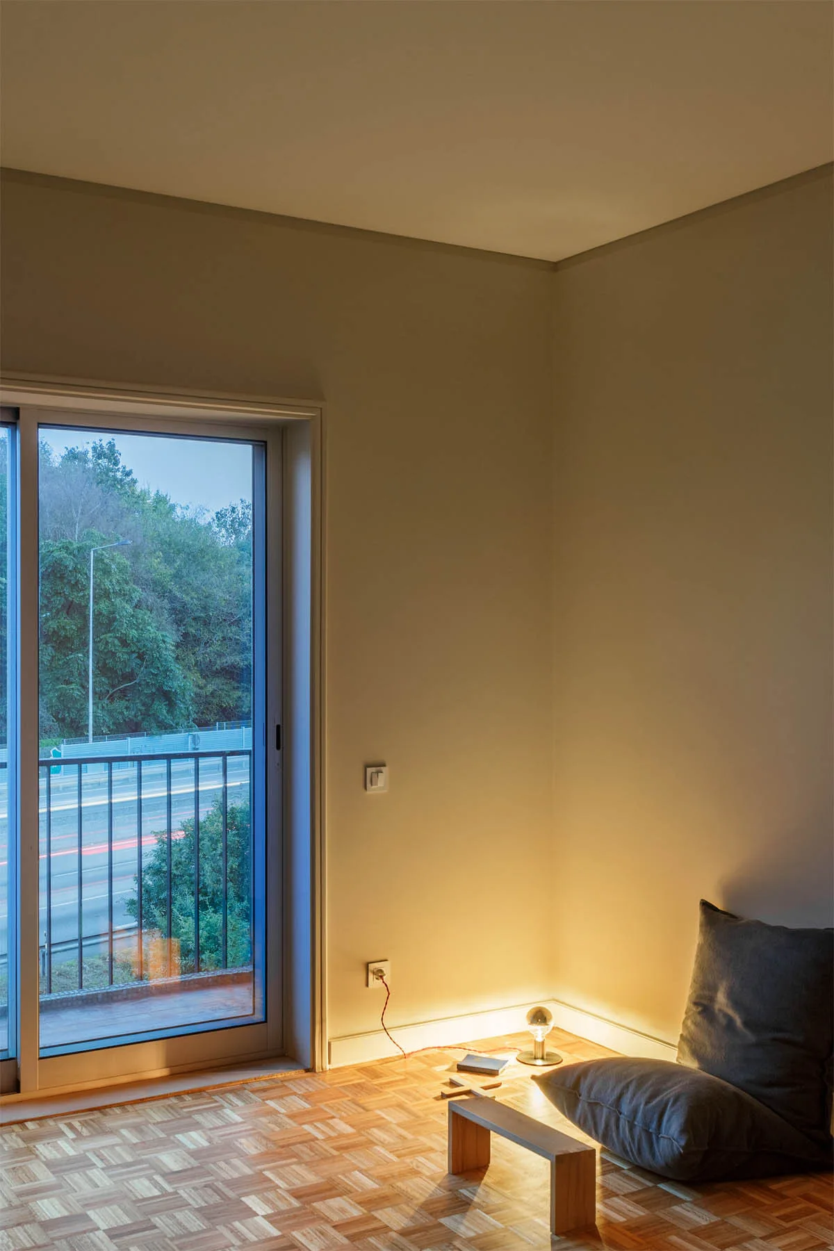 A minimalist corner featuring parquet wood flooring, gray floor cushions, a low wooden bench, and a small illuminated lamp against beige walls.