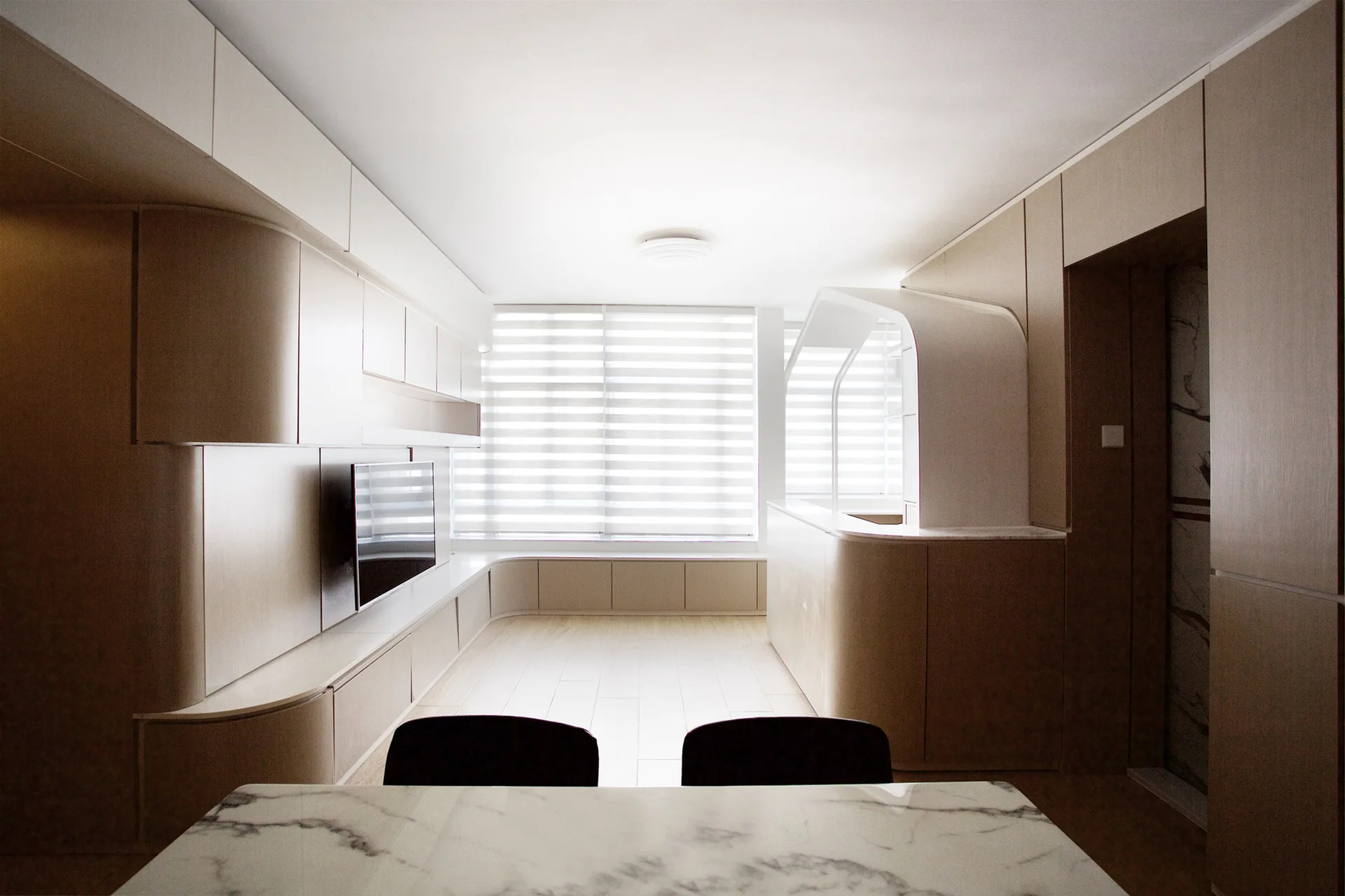 A minimalist room featuring seamless light wood cabinetry with curved corners, a large window with horizontal blinds, and a marble table in the foreground.