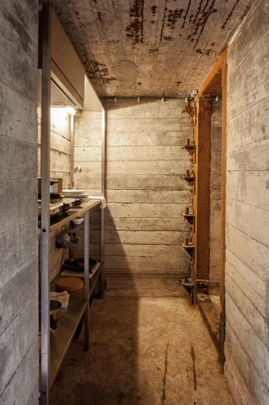 An industrial galley kitchen features stainless steel shelving with a sink and stovetop set against raw, board-marked concrete walls.