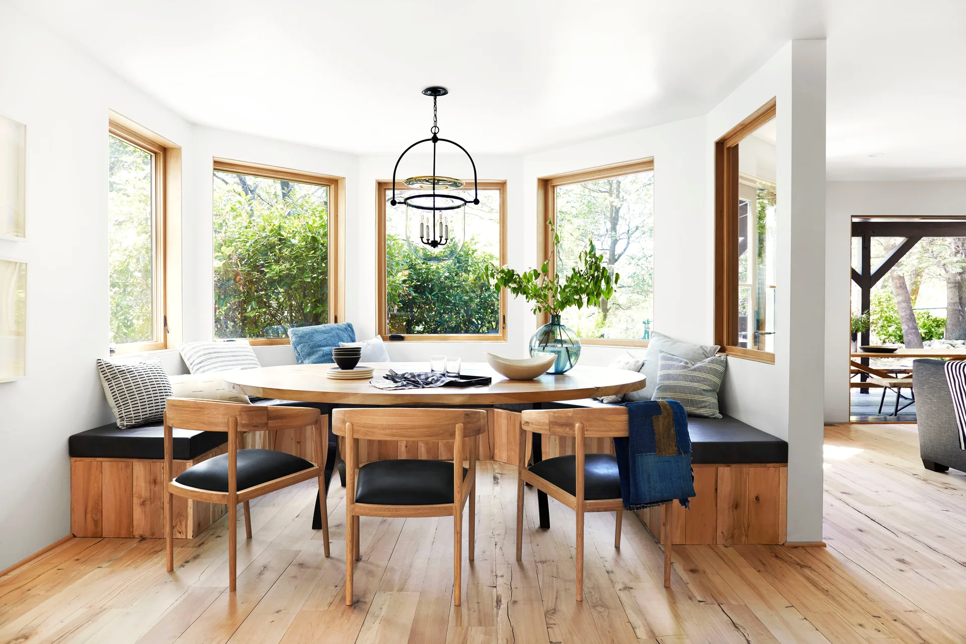 A sunlit breakfast nook features a curved wooden built-in banquette, a round dining table, wood chairs, and large bay windows.